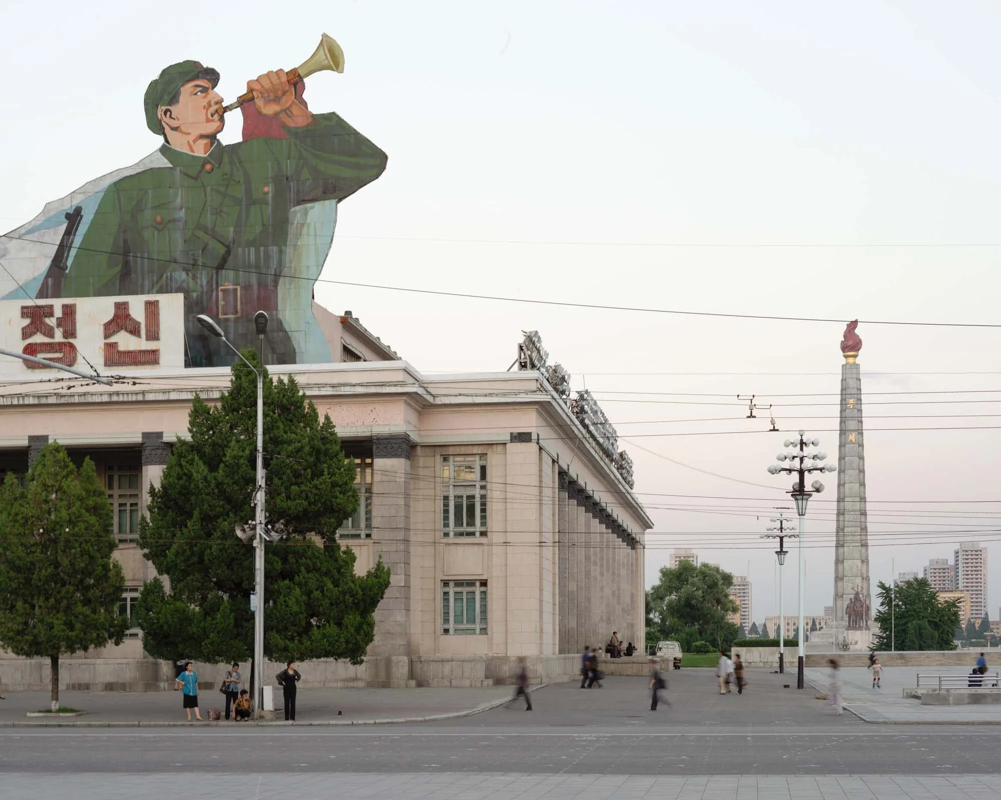 Propagandaplakat nahe dem Juche-Turm in Pjöngjang, Nordkorea: Ein großflächiges, zugeschnittenes Wandbild zeigt einen Soldaten in Uniform, der in ein Signalhorn bläst – ein klassisches Motiv der nordkoreanischen Mobilisierungspropaganda. Im Hintergru