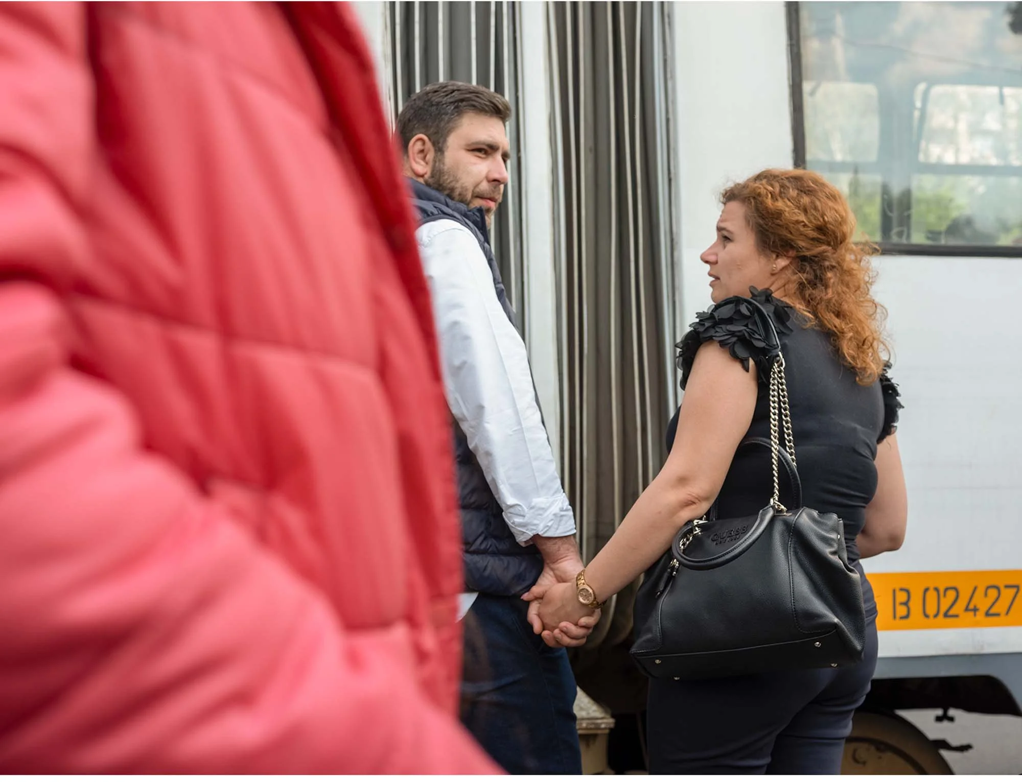 Ein Mann und eine Frau halten Händchen, während sie  auf dir Tram/ Strassenbahn in Bukarest/ Rumänien warten  warten  Im Auf der linken Seite sind die Schultern einer Person in einem roten Kleid oder Jacke sichtbar, die im Vordergrund steht.