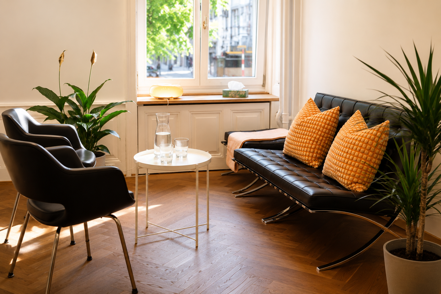 Gemütliches Wohnzimmer mit schwarzen Sesseln, einer schwarzen Ottomane mit orangenen Kissen, einem kleinen weißen Tisch mit Wasserkrügen und Gläsern, einer großen grünen Pflanze, einem Fenster mit Blick nach draußen, einem Nachttisch mit einer Lampe 