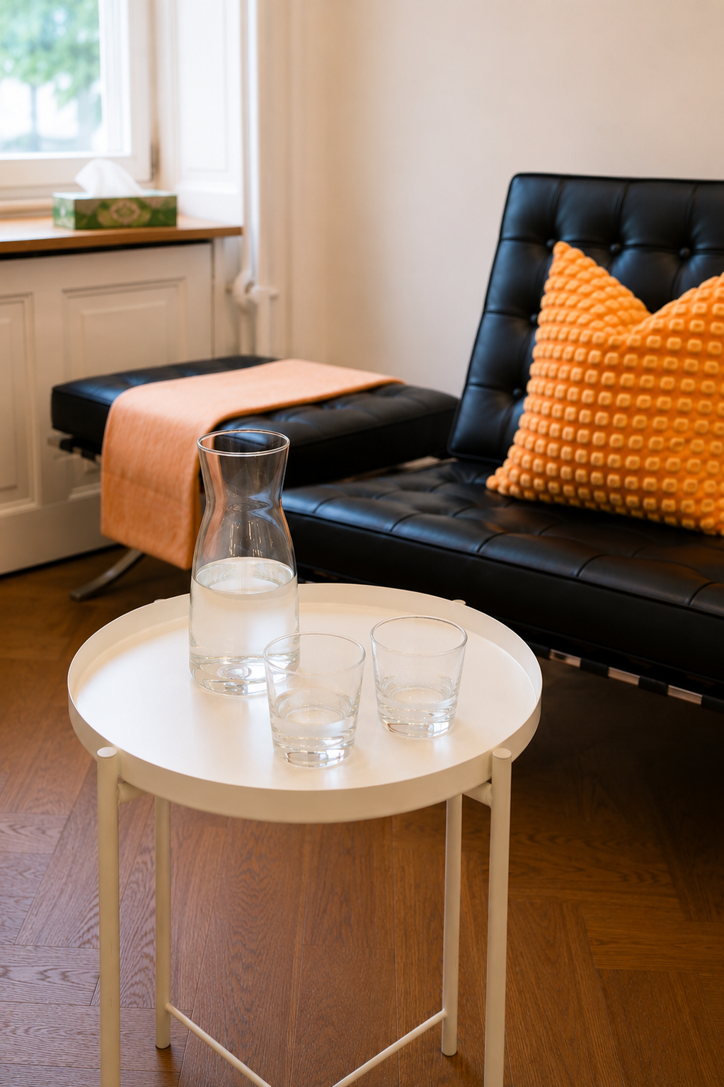 Ein Wohnzimmer mit einem schwarzen Ledersofa, einem orangefarbenen Kissen, einem weißen Beistelltisch mit Wasserkrug und Gläsern, einem Fenster mit Vorhängen, einem Heizkörper, einer beige Decke auf einer Bank, und einem Karton-Box auf dem Fensterbre