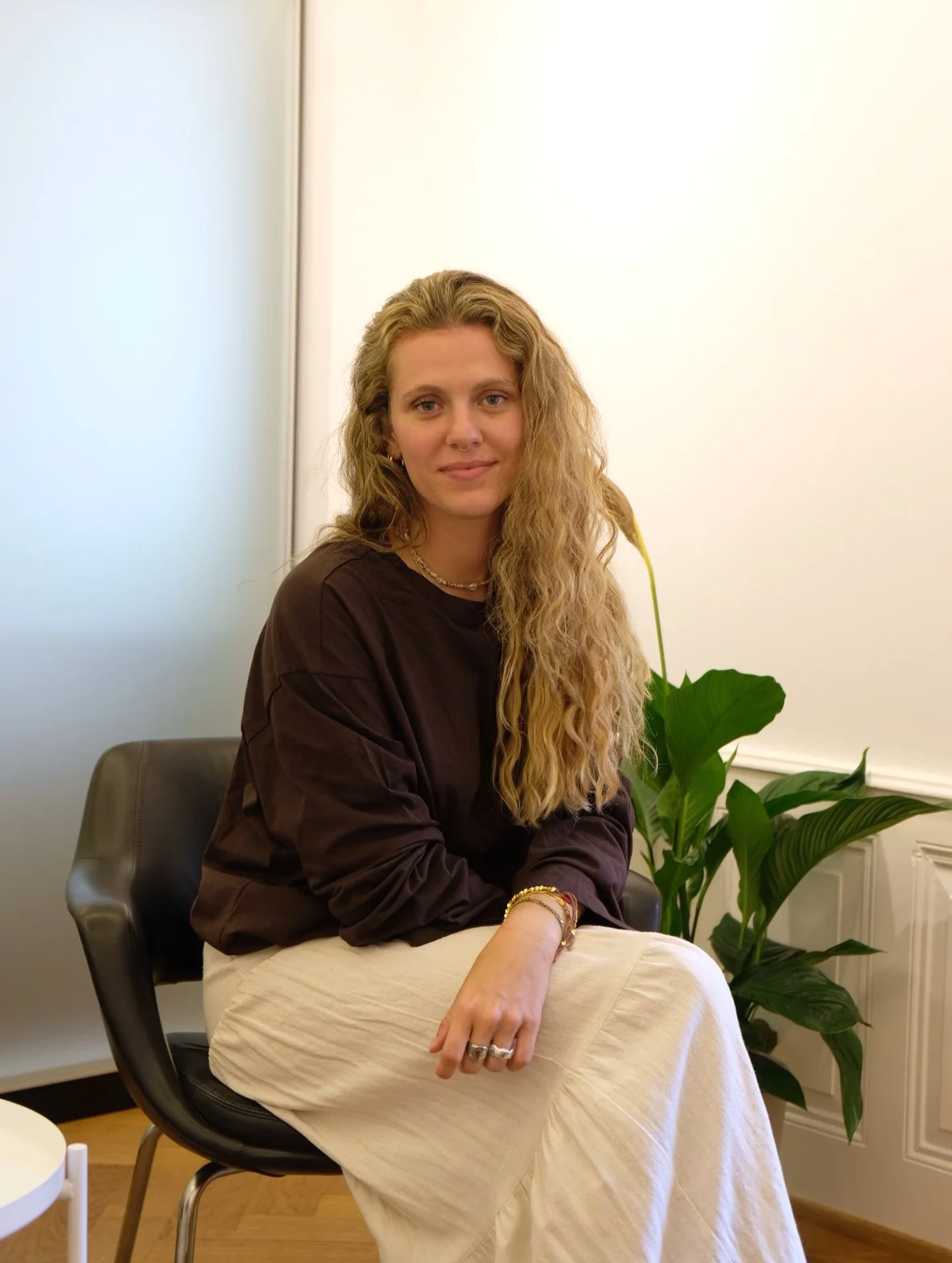 Eine junge Frau mit langen, lockigen blonden Haaren sitzt in einem Raum mit weißen Wänden und einer großen grünen Pflanze. Sie trägt dunkle Bluse und einen weißen Rock sowie Schmuck und lächelt leicht.