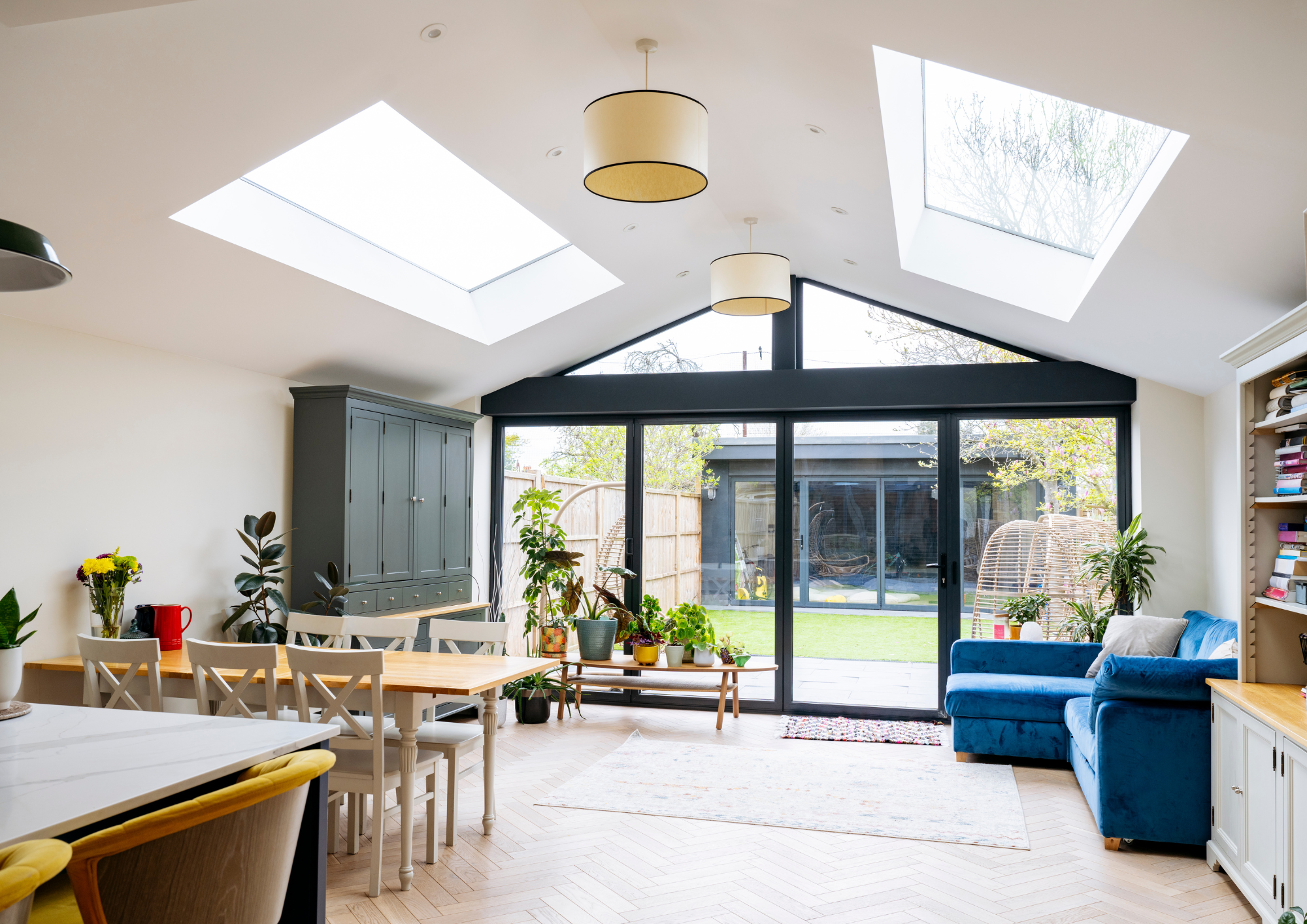 Living room with large skylights, plants, blue sofa, white dining table, and sliding doors to outside yard.