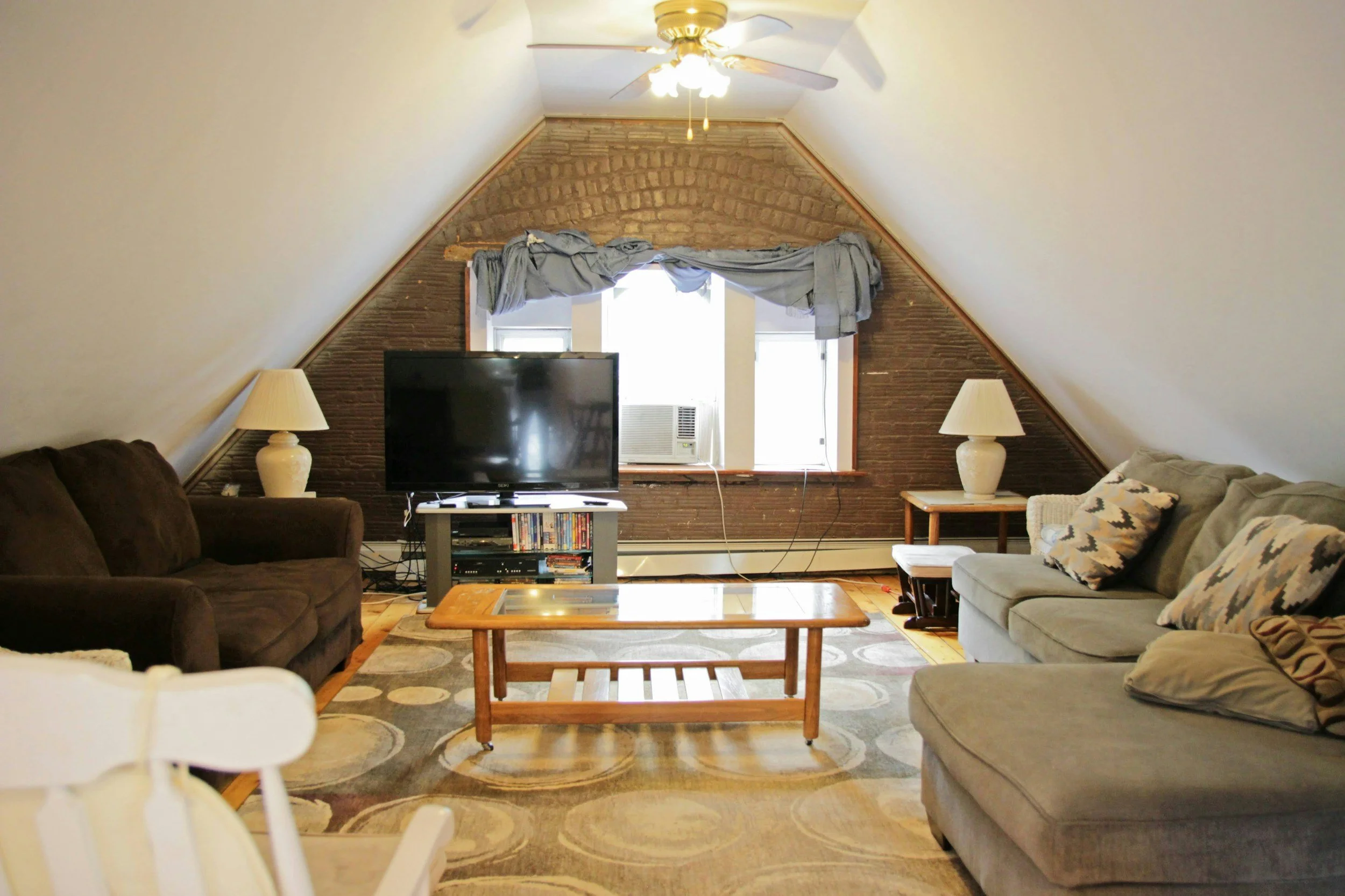 Living room with gabled ceiling, brick accent wall, window with curtains, television, ceiling fan, sofas, lamps, coffee table, and patterned area rug.