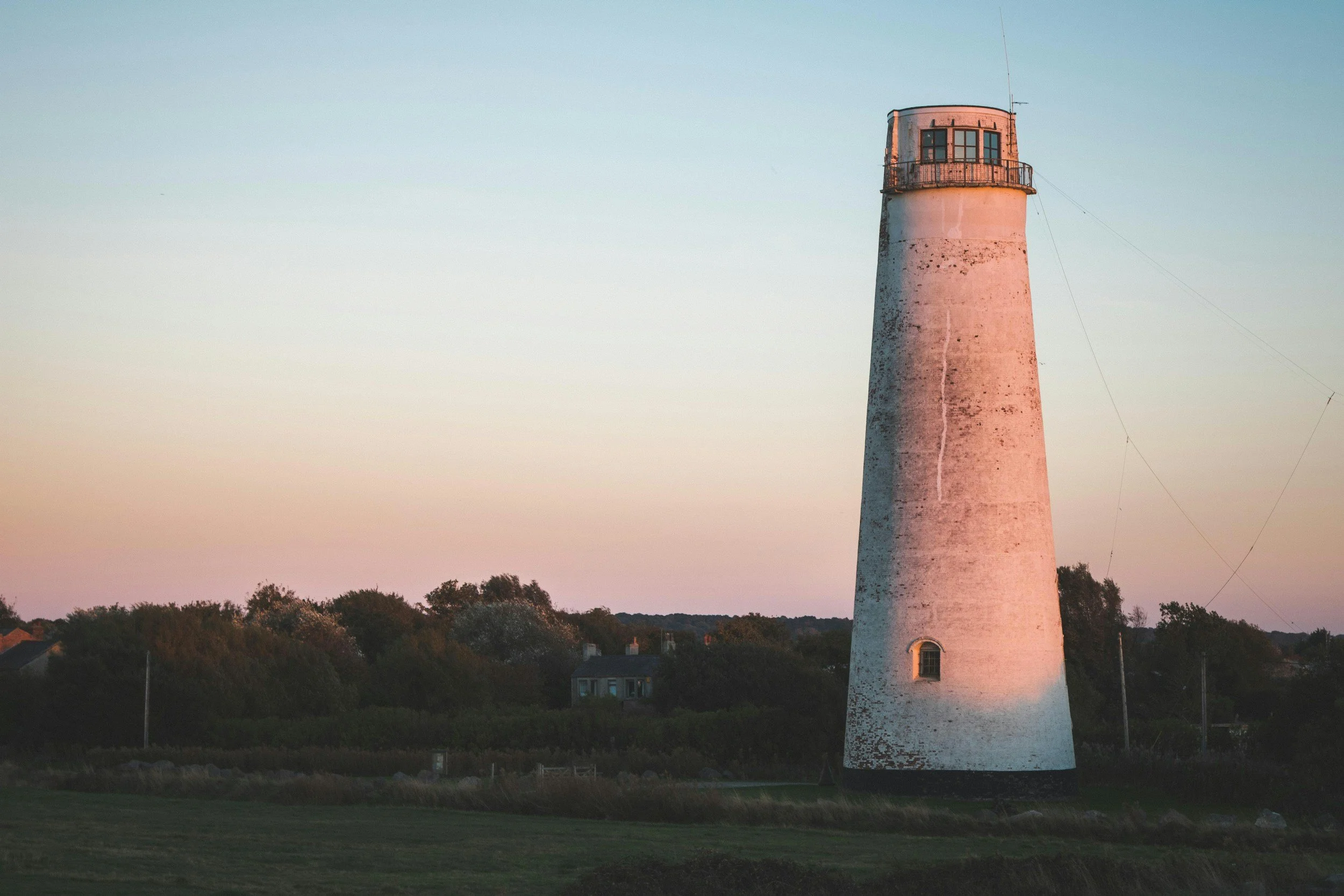 A tall, white lighthouse with a rounded top and small windows, set against a sunset sky with soft pastel colors, surrounded by trees and a few houses.