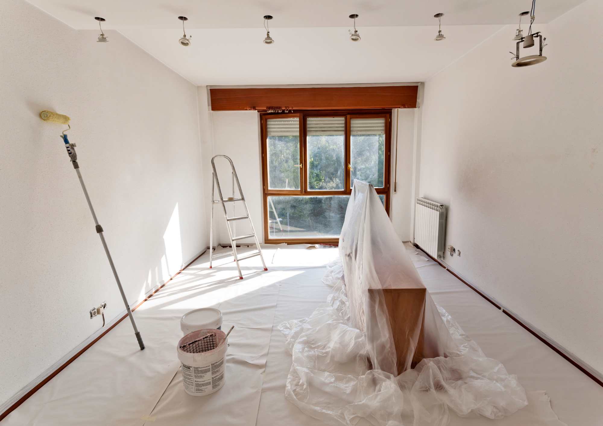 A room under renovation with white walls, a large window, a ladder, and a paint roller. Painting supplies, including a bucket of paint and plastic sheets, are on the floor.