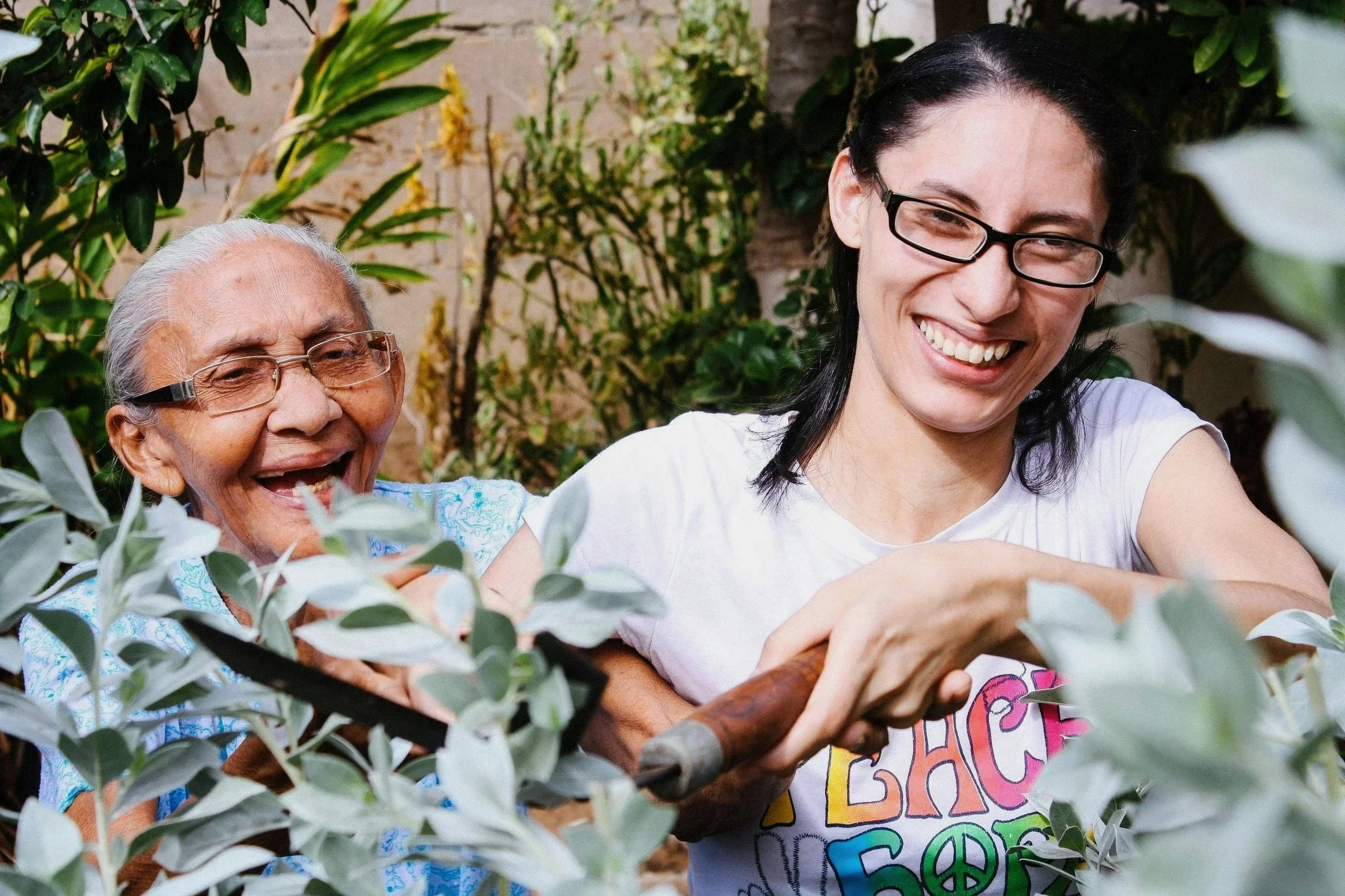 Two women, a senior woman with gray hair and a younger woman with black hair and glasses, are smiling and enjoying time together outdoors amid green plants, possibly in a garden or park.