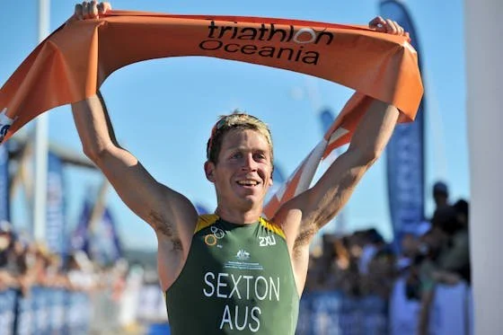 Athlete in a green sports uniform holding an orange towel above his head in a finish line area at a triathlon event, with a crowd and banners in the background.