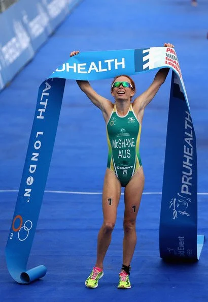 Female athlete crossing the finish line at a triathlon, holding a blue finish line banner, wearing a green and blue tri suit with Australian flag and sunglasses.