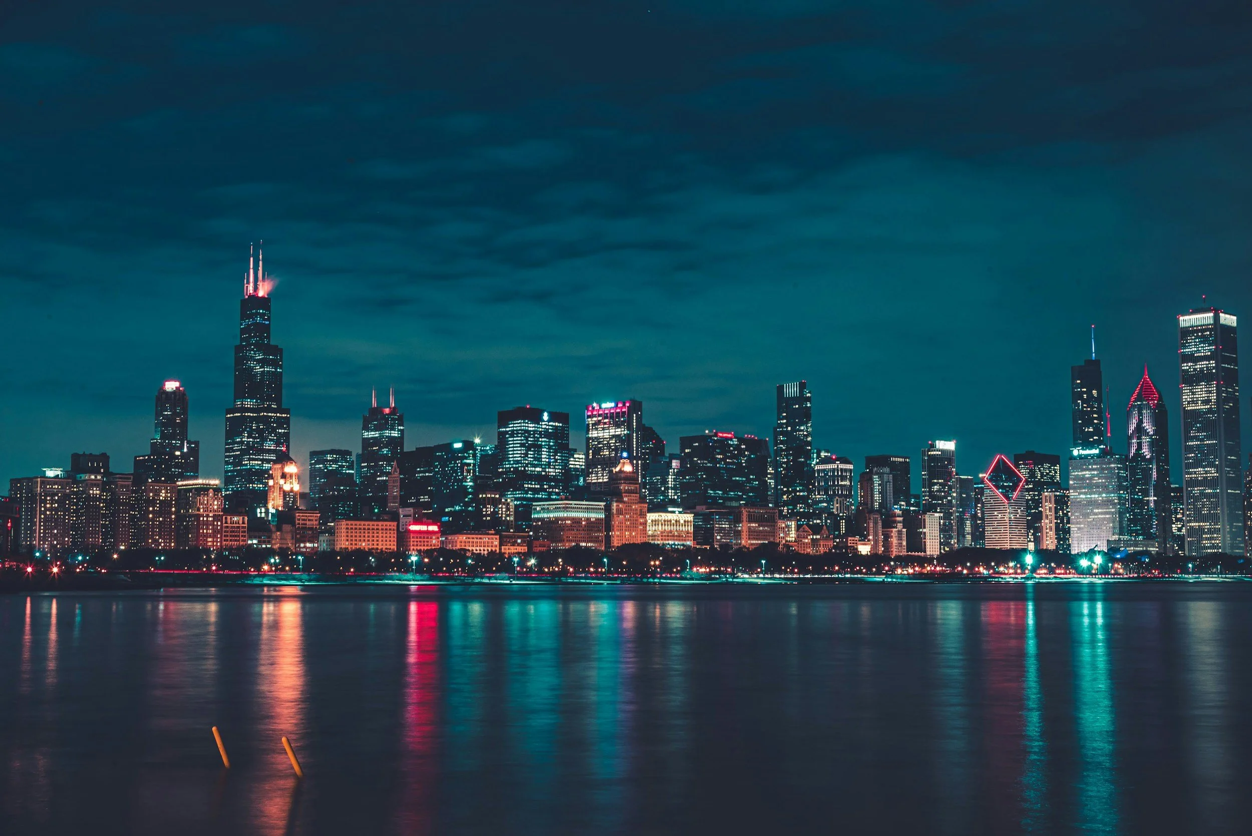 Nighttime city skyline with illuminated skyscrapers reflecting on a river, dark cloudy sky overhead.