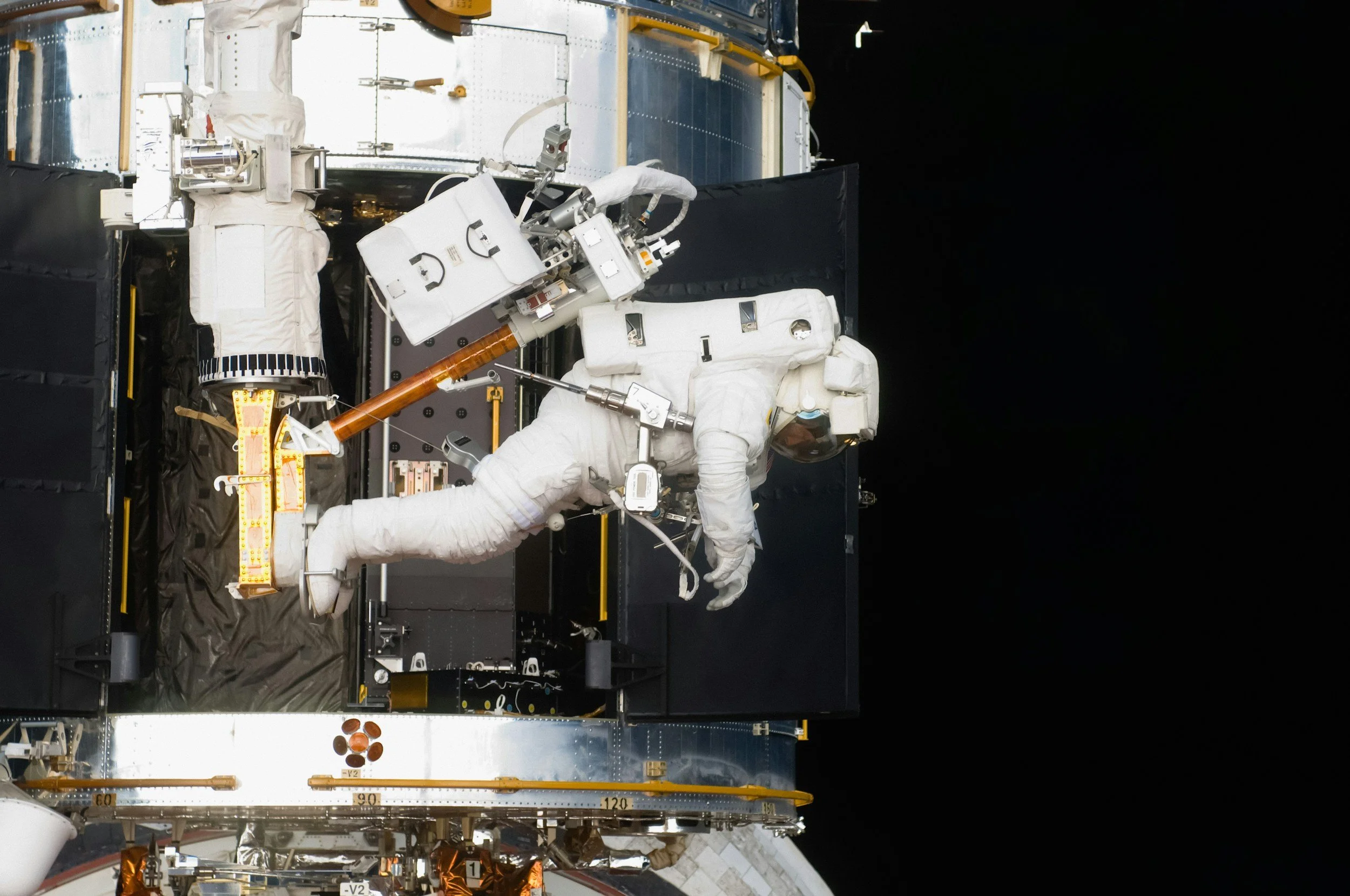 An astronaut in a white suit and helmet working outside the International Space Station in space, with a black background and parts of the space station visible.