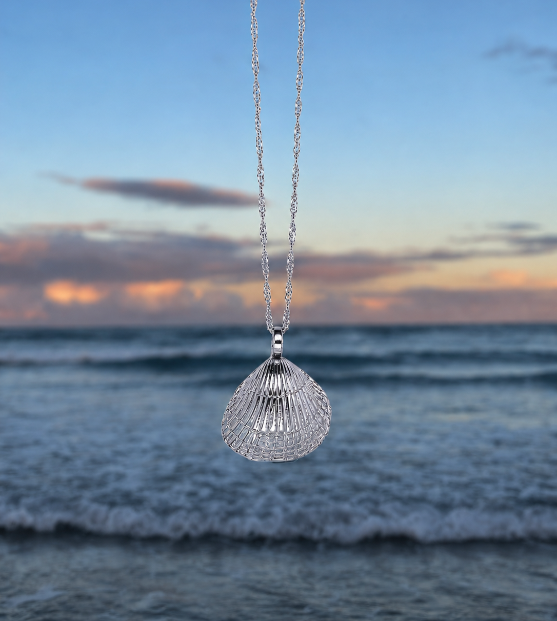 A silver necklace with a shell pendant hanging in front of a blurred beach and ocean at sunset.