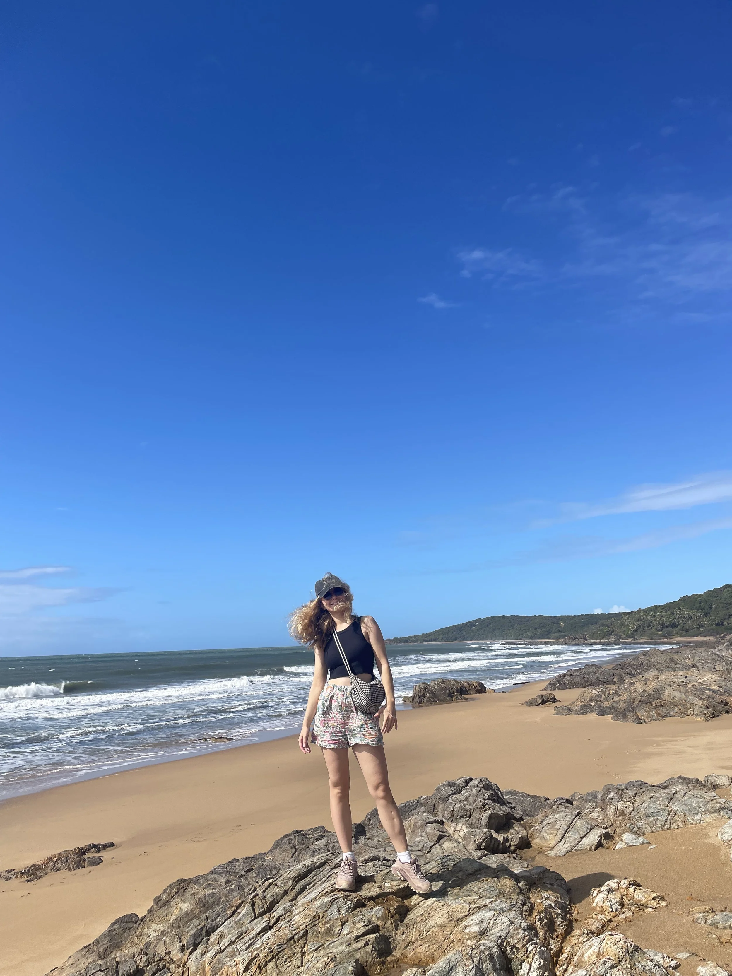 A woman standing on rocks at a beach with ocean waves, sandy shore, and clear blue sky in the background.