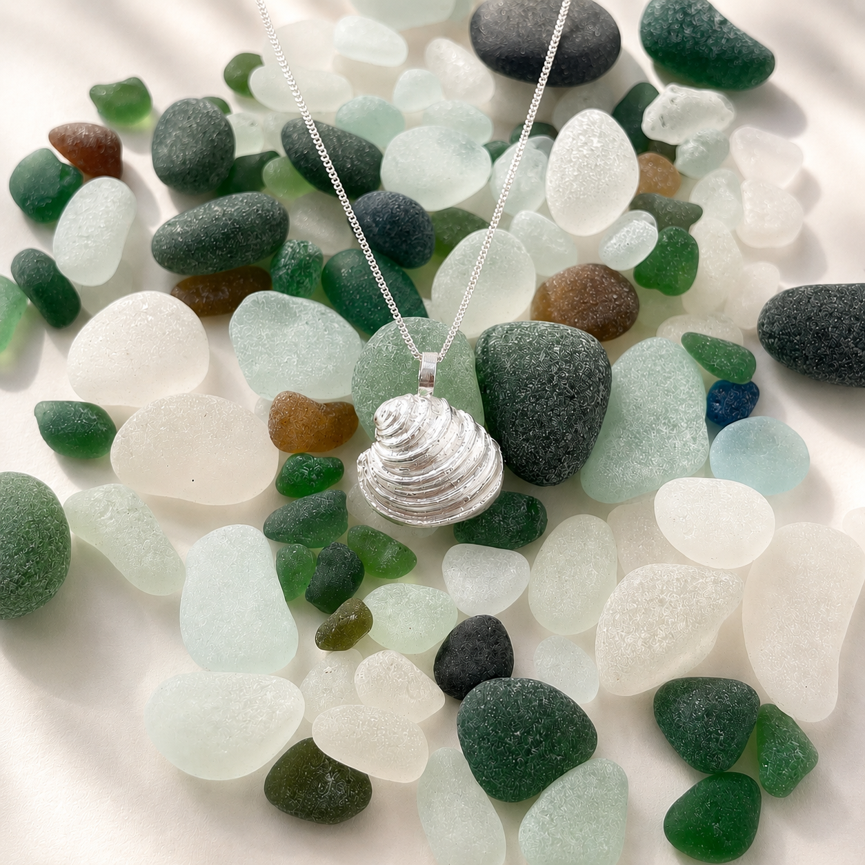 Silver seashell necklace on a silver chain among colorful sea glass pieces in white, green, clear, black, and brown tones.