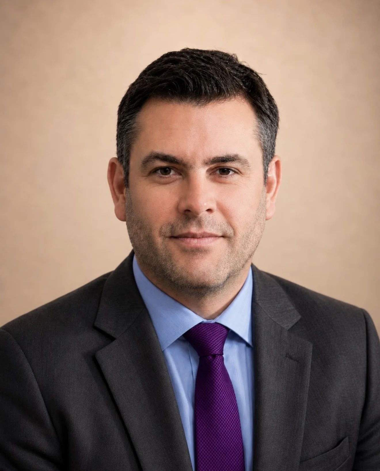 A professional headshot of a man with dark brown hair, smiling, dressed in a dark gray suit, light blue shirt, and purple tie, against a beige background.