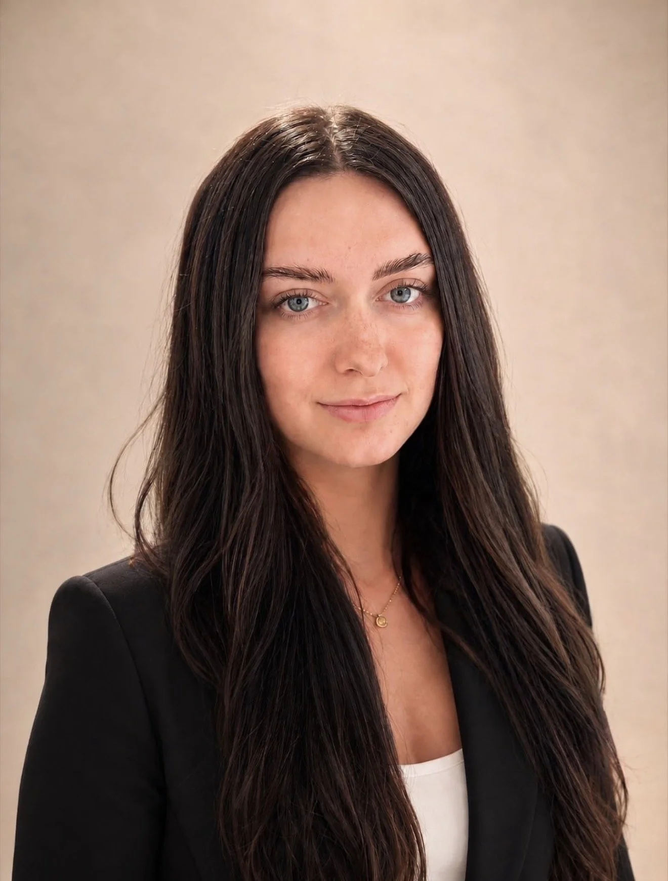 A woman with long dark hair and blue eyes wearing a black blazer, white top, and a small gold necklace.