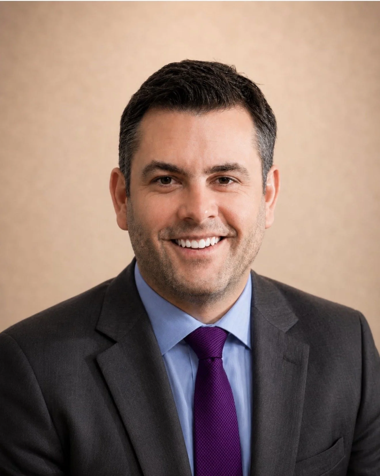Professional portrait of a smiling man with dark hair in a suit, blue shirt, purple tie, and a beige background.