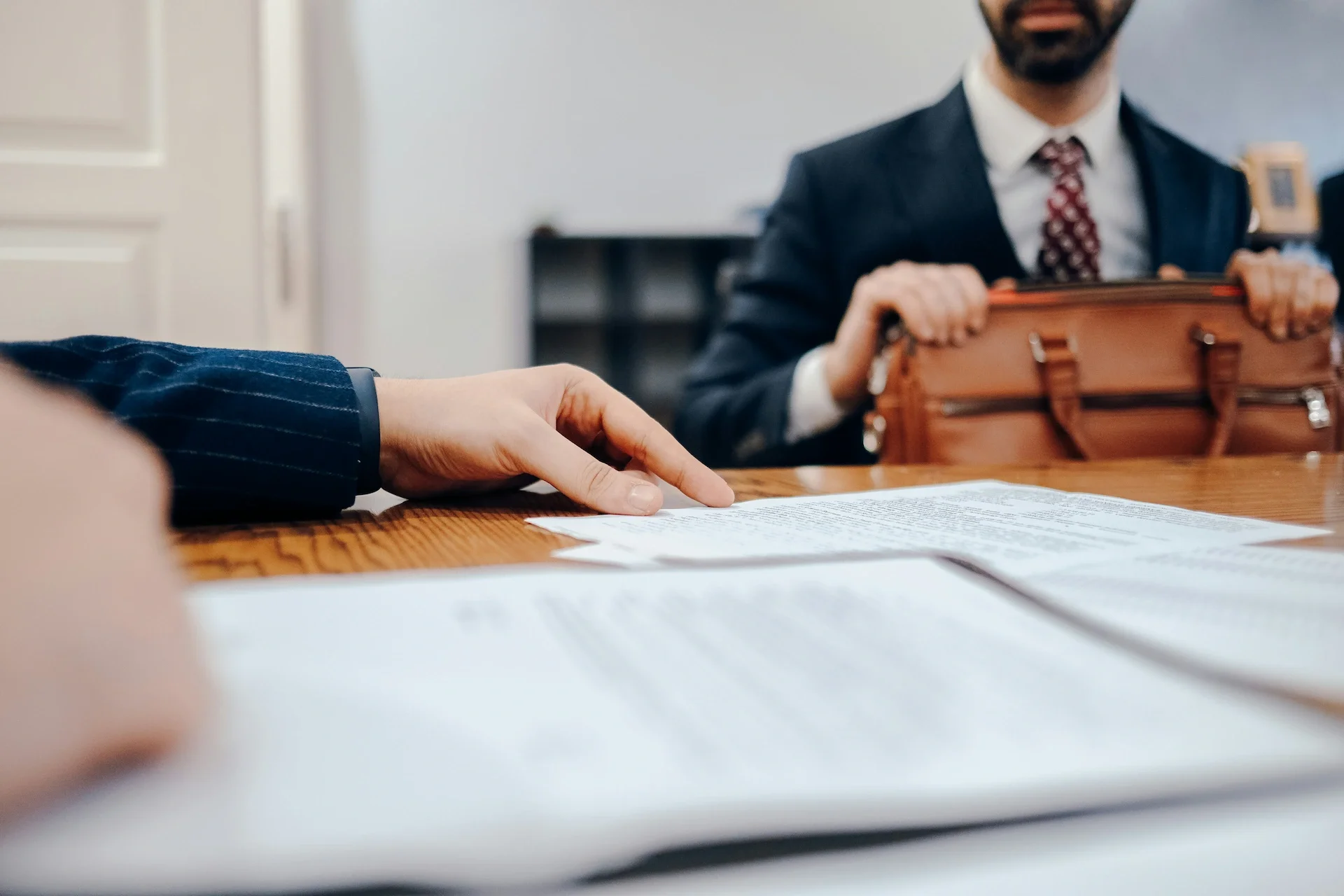 Close-up of a person's hand pointing at a document on a wooden table during a business meeting, with another person in a suit in the background holding a brown leather briefcase.