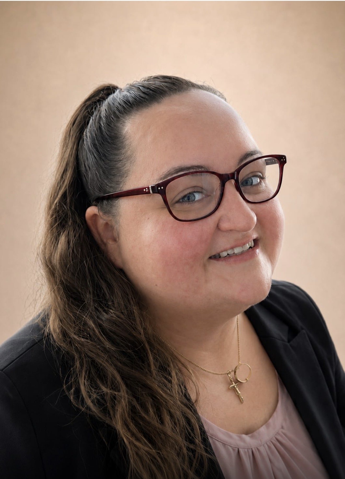 A woman with long brown hair, wearing glasses, smiling and looking to the side, dressed in a black blazer with a pale pink top, and wearing jewelry including earrings and a necklace with a cross pendant, against a plain beige background.