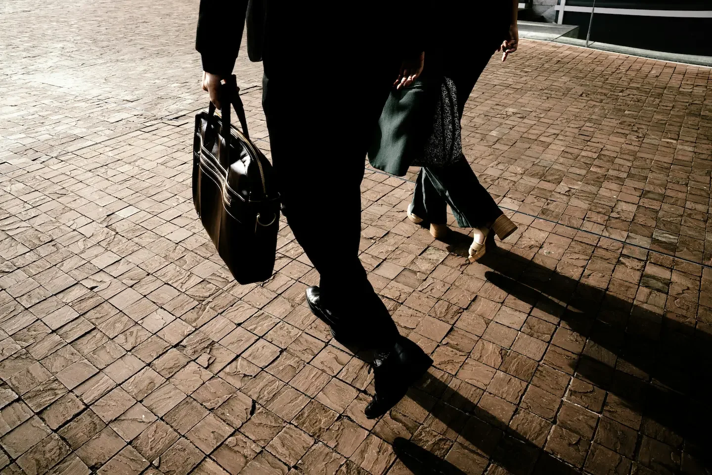 People walking on a brick sidewalk, one person dressed in black carrying a black handbag, and another person wearing high-heeled shoes.