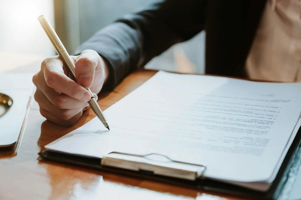 A person in business attire writing on a clipboard with a silver pen on a wooden desk.