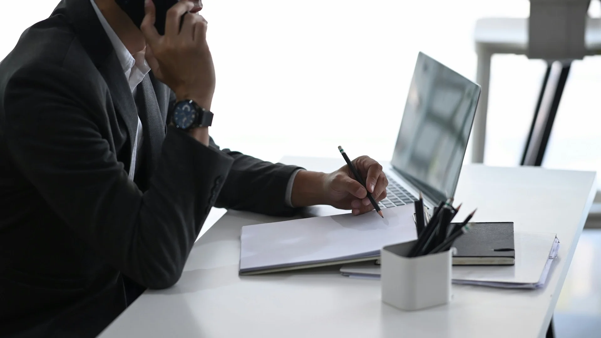 A person in business attire sitting at a white desk, talking on the phone, holding a pencil, with a laptop and office supplies nearby.