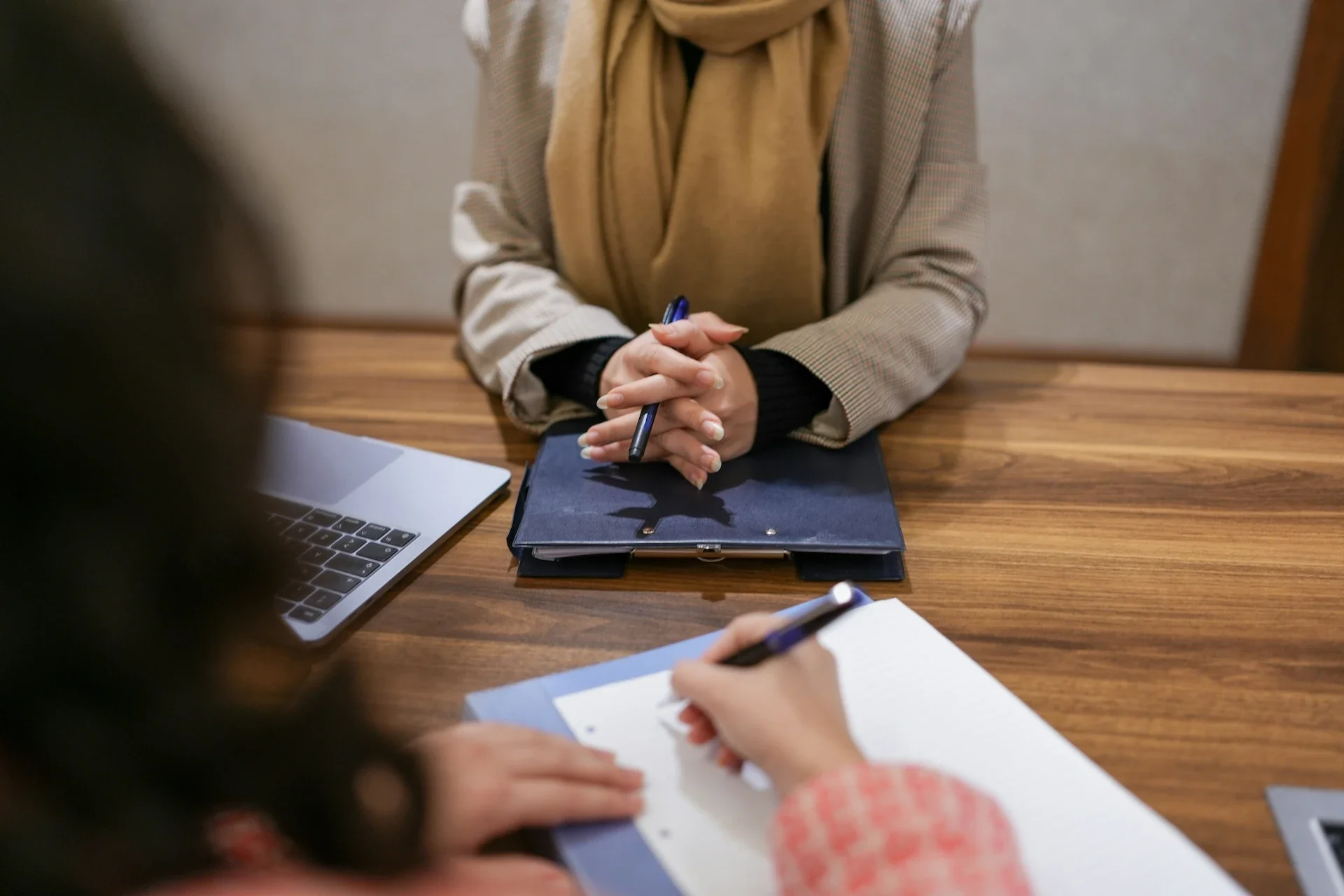 A person in a checkered blazer and beige scarf sits at a wooden table with a closed laptop, a blue notebook, and a pen. Another person is taking notes in a notebook.