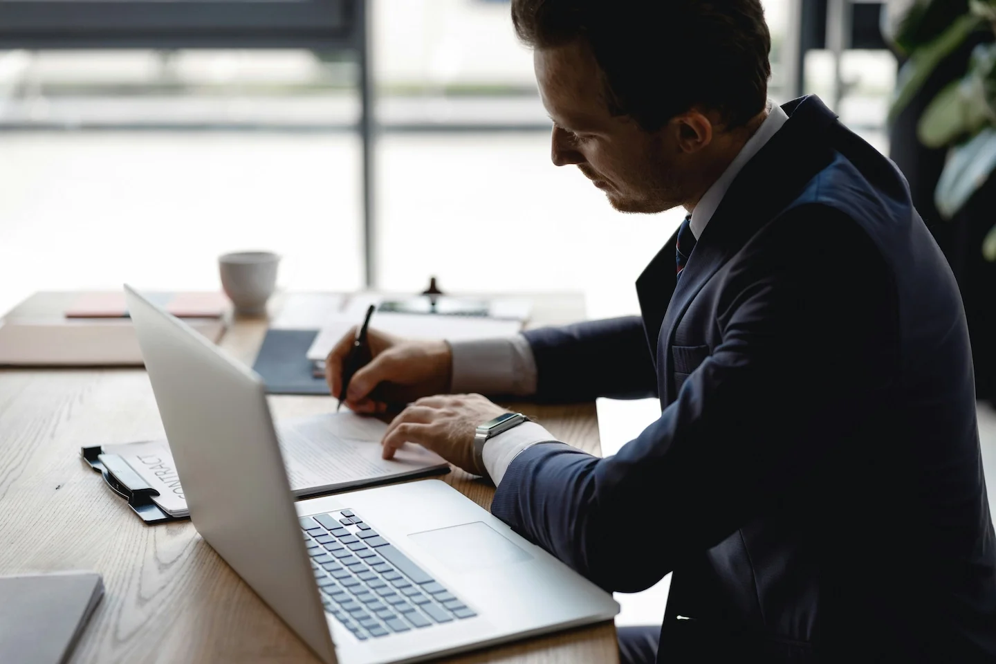 Businessman sitting at a desk, writing on documents, with a laptop open in front of him in an office setting.