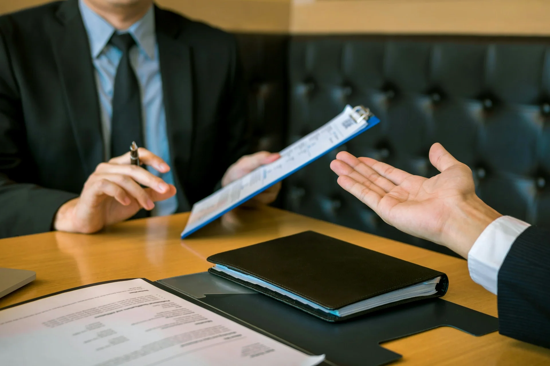 A person in a black suit and tie is holding a clipboard and writing notes, while another person in a business suit gestures with their hand during a meeting at a wooden table with documents and a black folder.