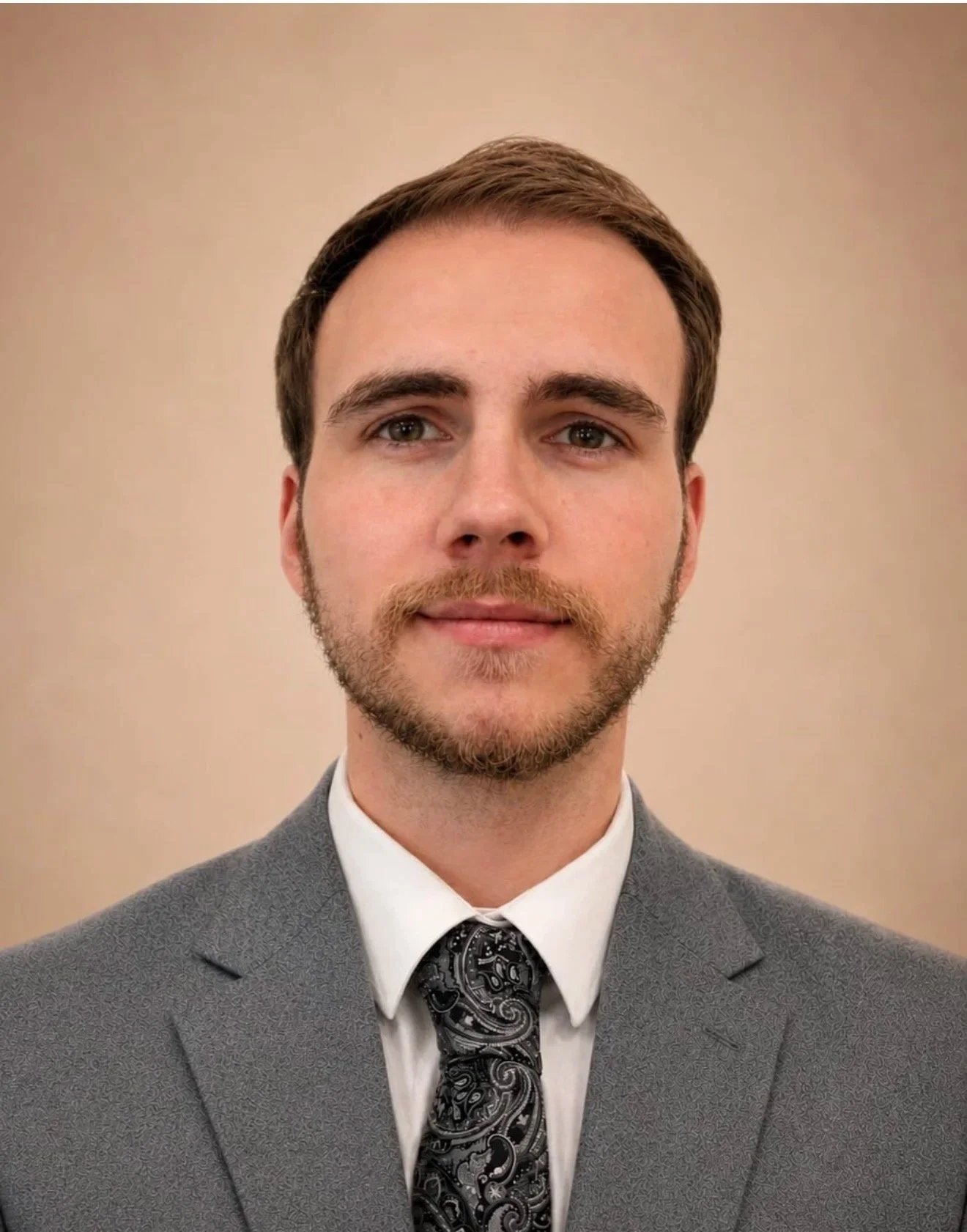 A young man with light skin, brown hair, and a beard wearing a gray suit, white shirt, and a paisley patterned tie, standing against a beige background.