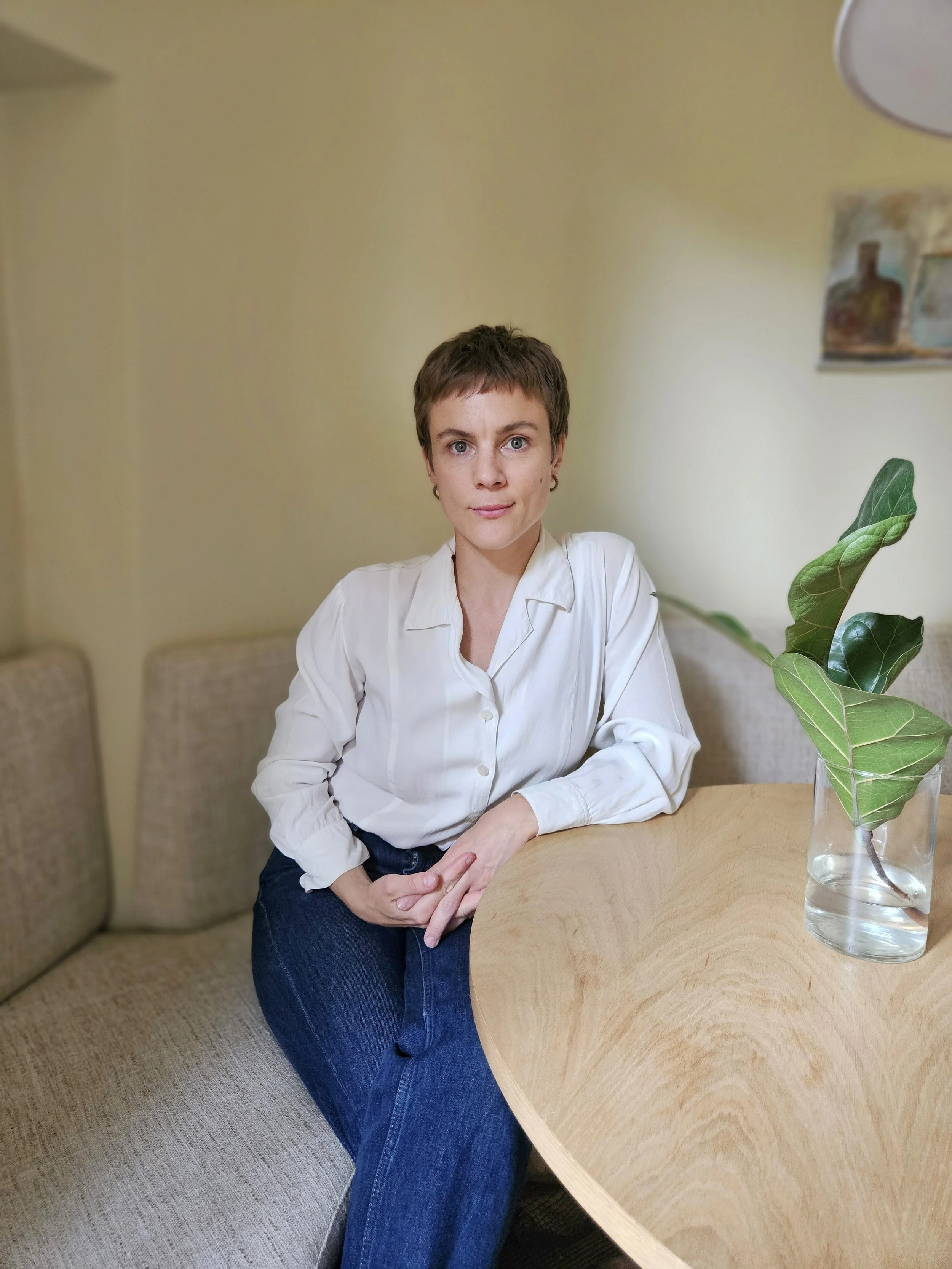 Psykolog som bedriver psykodynamisk terapi för ångest depression trauma och stress, vit blus och blå jeans sitter vid ett runt bord i ett rum med beige väggar. På bordet finns en vas med gröna blad.