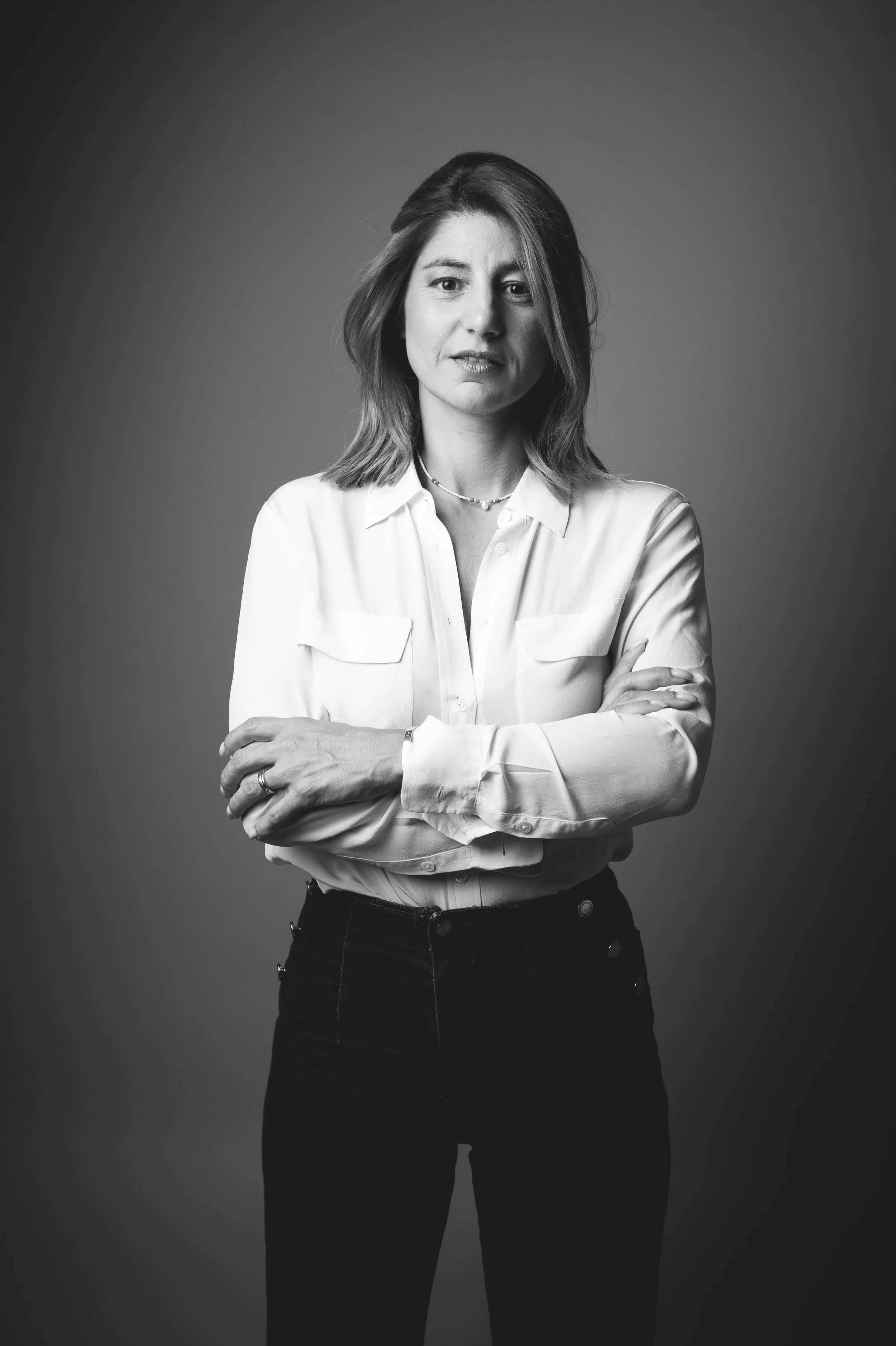 Portrait en noir et blanc d'une femme avec des cheveux mi-longs, portant une chemise blanche à manches longues, un collier et des bracelets, avec les bras croisés.