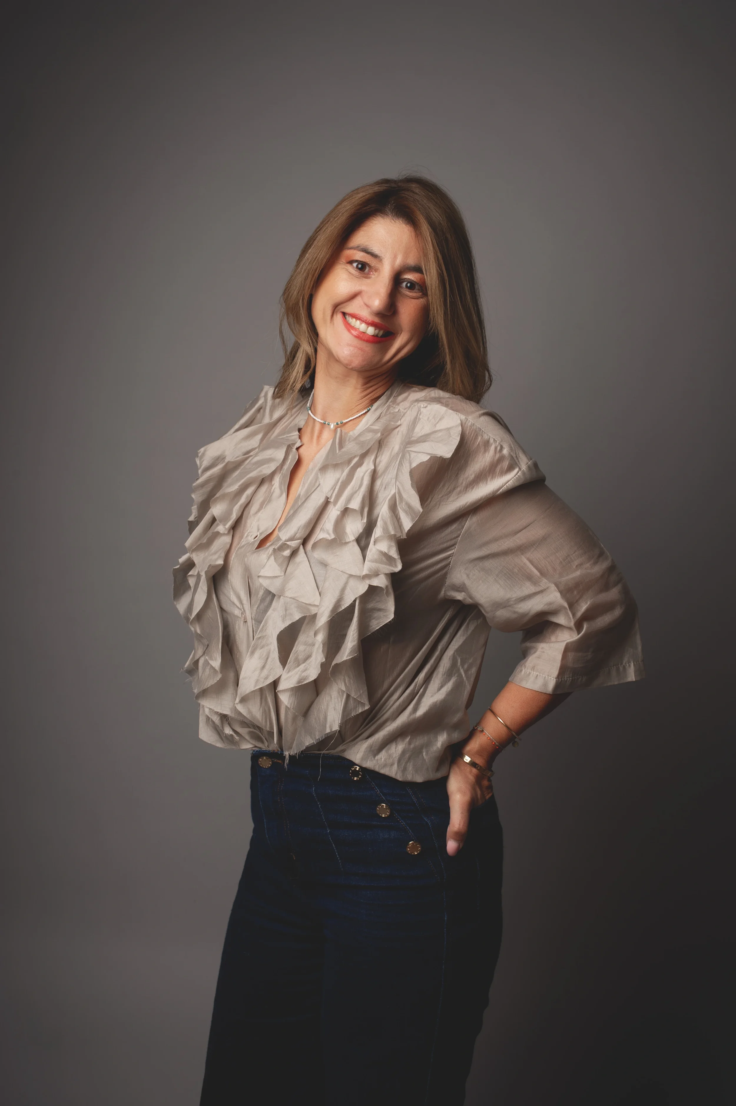 Jeune femme souriante posant avec une main sur la hanche, portant un haut en tissu beige avec des volants et un collier.