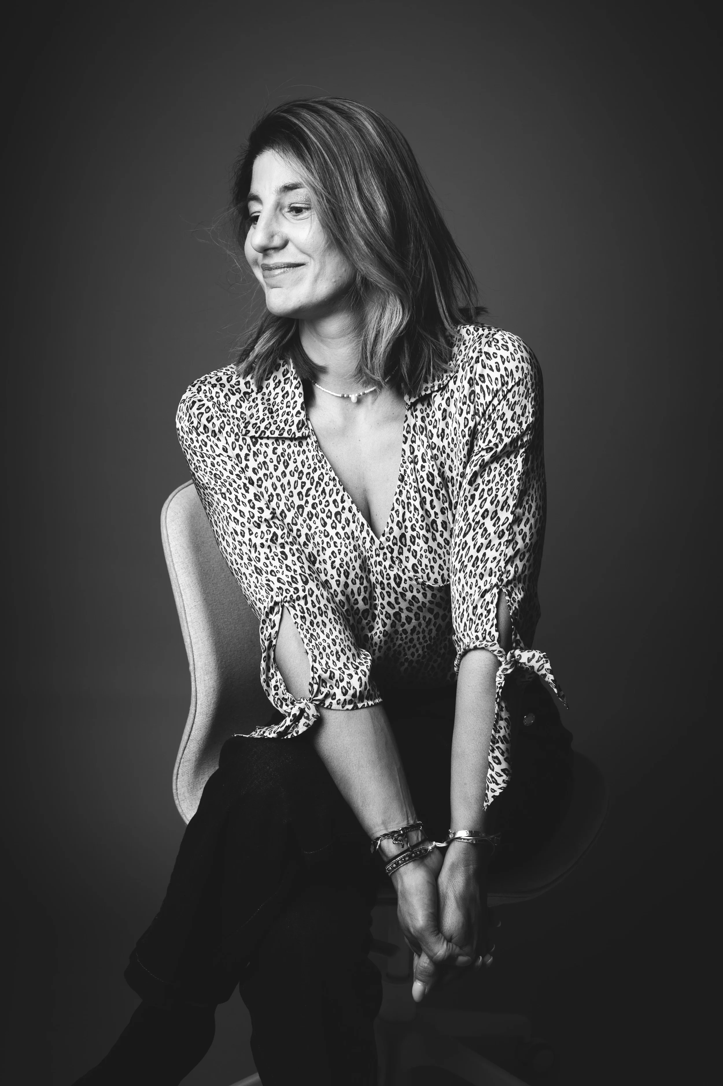 Une femme aux cheveux longs et ondulés, portant un chemisier à imprimé léopard, assise sur une chaise, dans un studio avec fond gris. La photo est en noir et blanc.