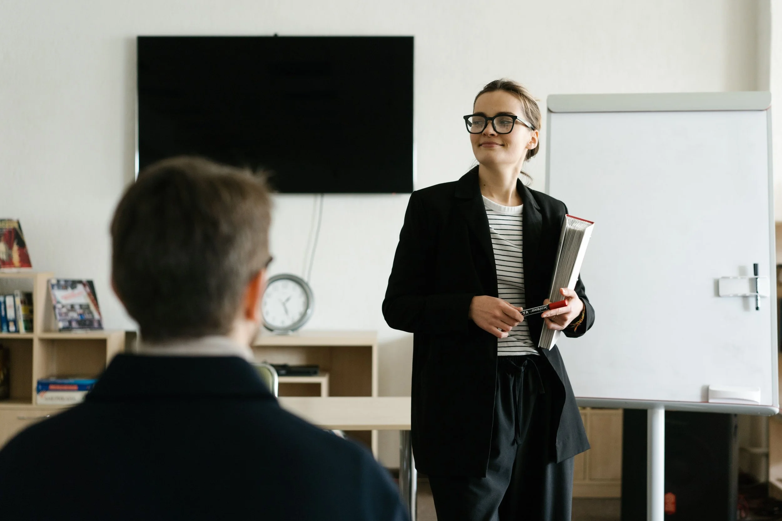 Une femme souriante portant des lunettes noires, une veste noire, et tenant des livres, se trouve dans une salle de classe en train de parler à un groupe.