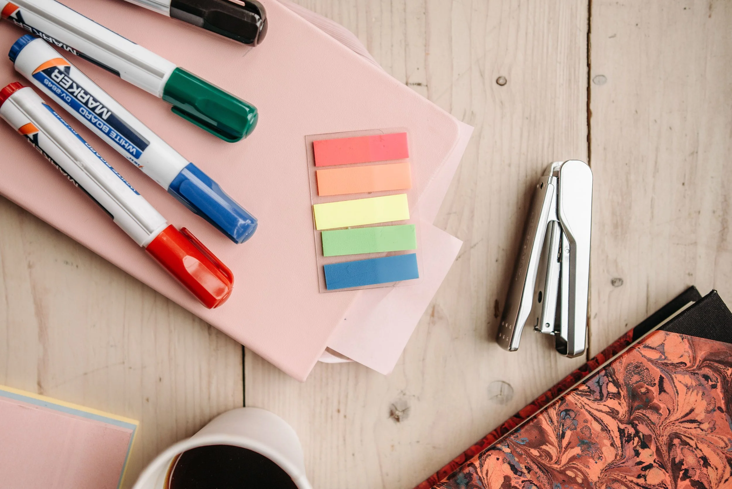 Une table en bois avec des marqueurs de différentes couleurs, des blocs de post-it, une perforatrice, une tasse de café, et un livre. Sur la table, il y a aussi un cahier rose clair et un autre cahier avec un motif rouge et noir.