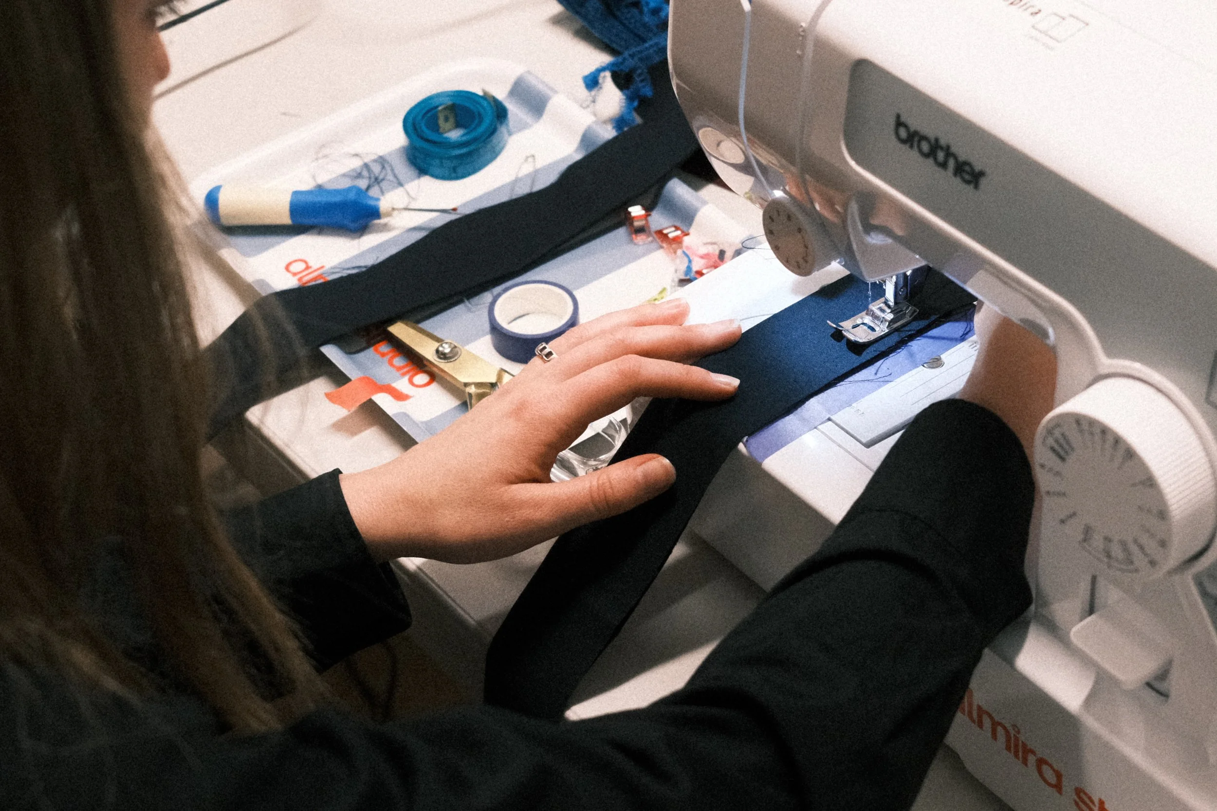 A person using a sewing machine on fabric, with sewing tools and measurement tape nearby, on a table with a paper or instruction sheet underneath.