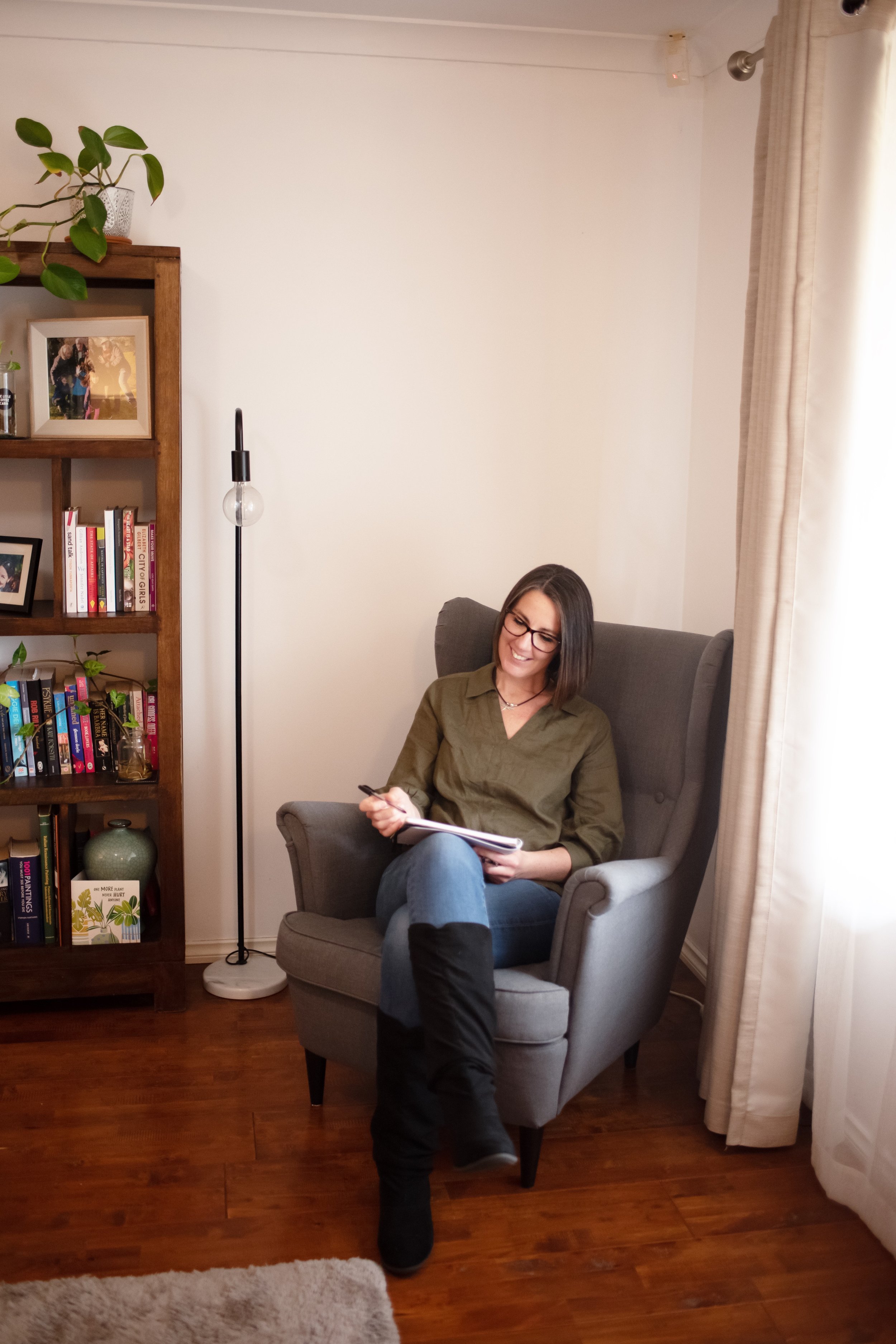 A woman with glasses and shoulder-length brown hair sitting in a gray armchair in a living room, holding a notebook and pen, smiling. To her left is a wooden bookshelf filled with books and decoration, and behind her is a tall floor lamp, a window with curtains, and a plain white wall.