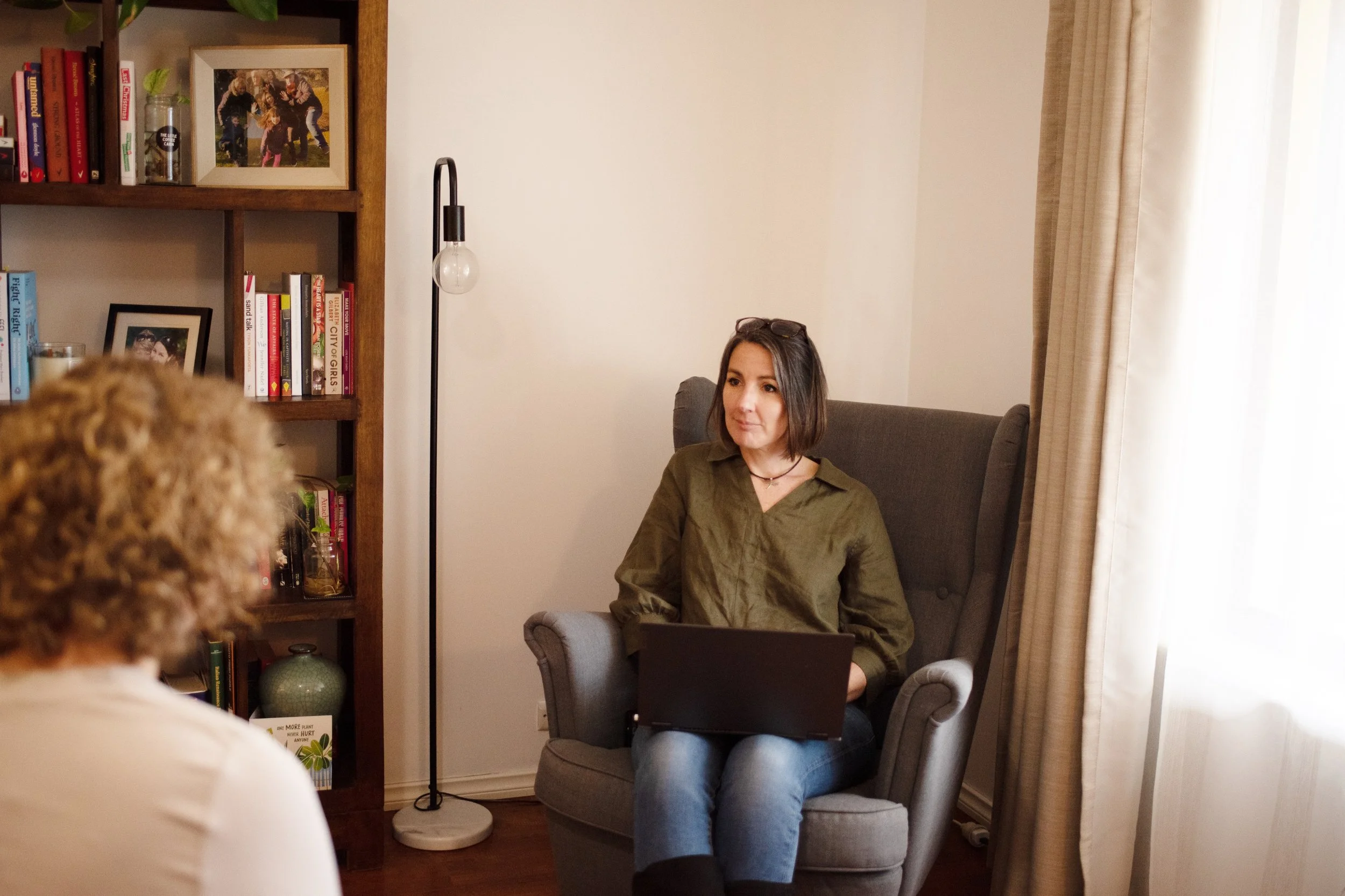 A woman with shoulder-length brown hair sitting in a gray upholstered armchair in a living room, wearing a green shirt and glasses on her head, looking to her right. Behind her is a beige curtain, a white wall, a floor lamp, and a wooden bookshelf filled with books and framed photographs.