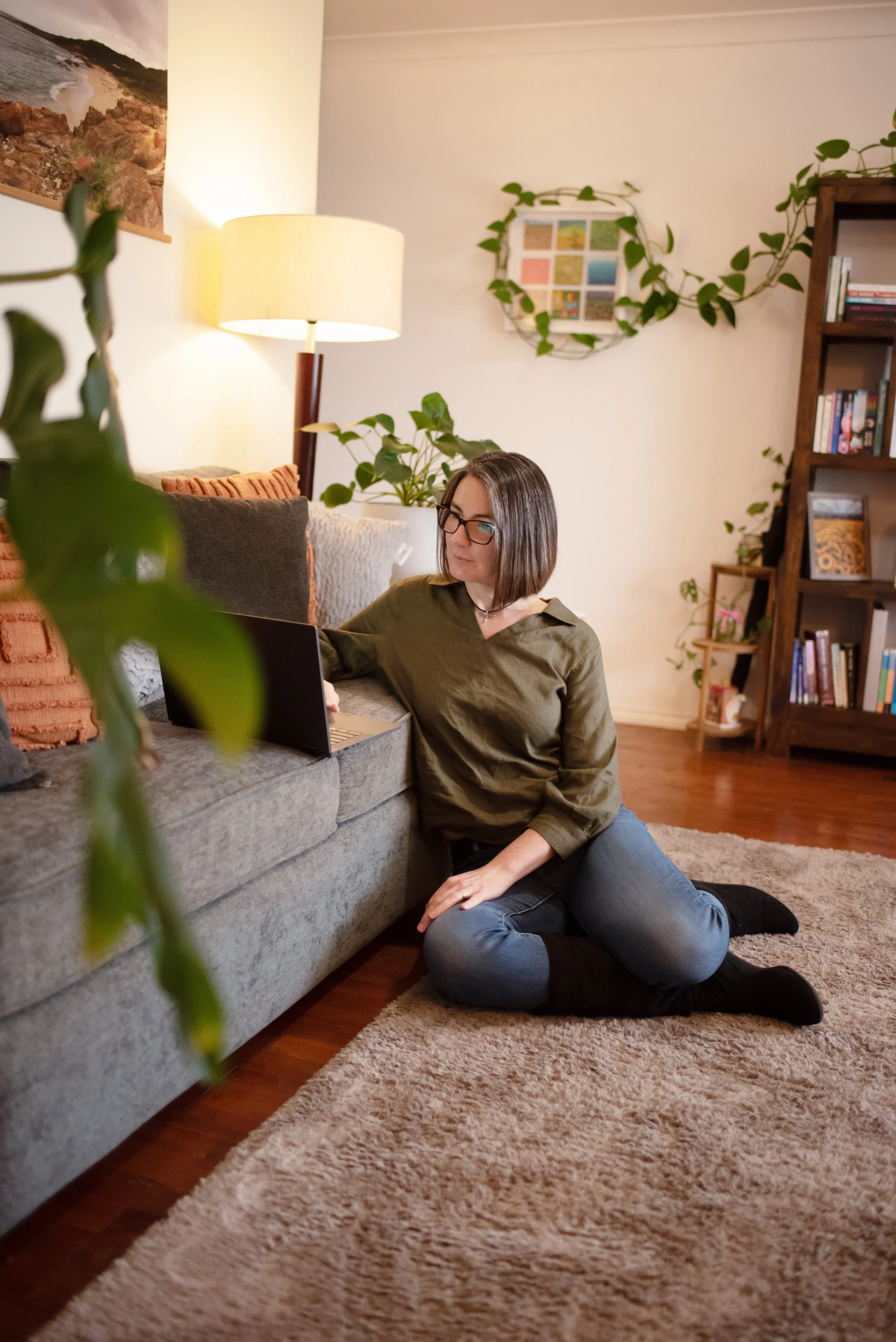 A woman with glasses and shoulder-length brown hair is sitting on a carpeted floor, leaning against a couch while working on a laptop. She is wearing a green shirt and black socks, with her legs bent and one arm resting on the sofa. The room has bookshelves, plants, a lamp, and framed artwork on the wall.