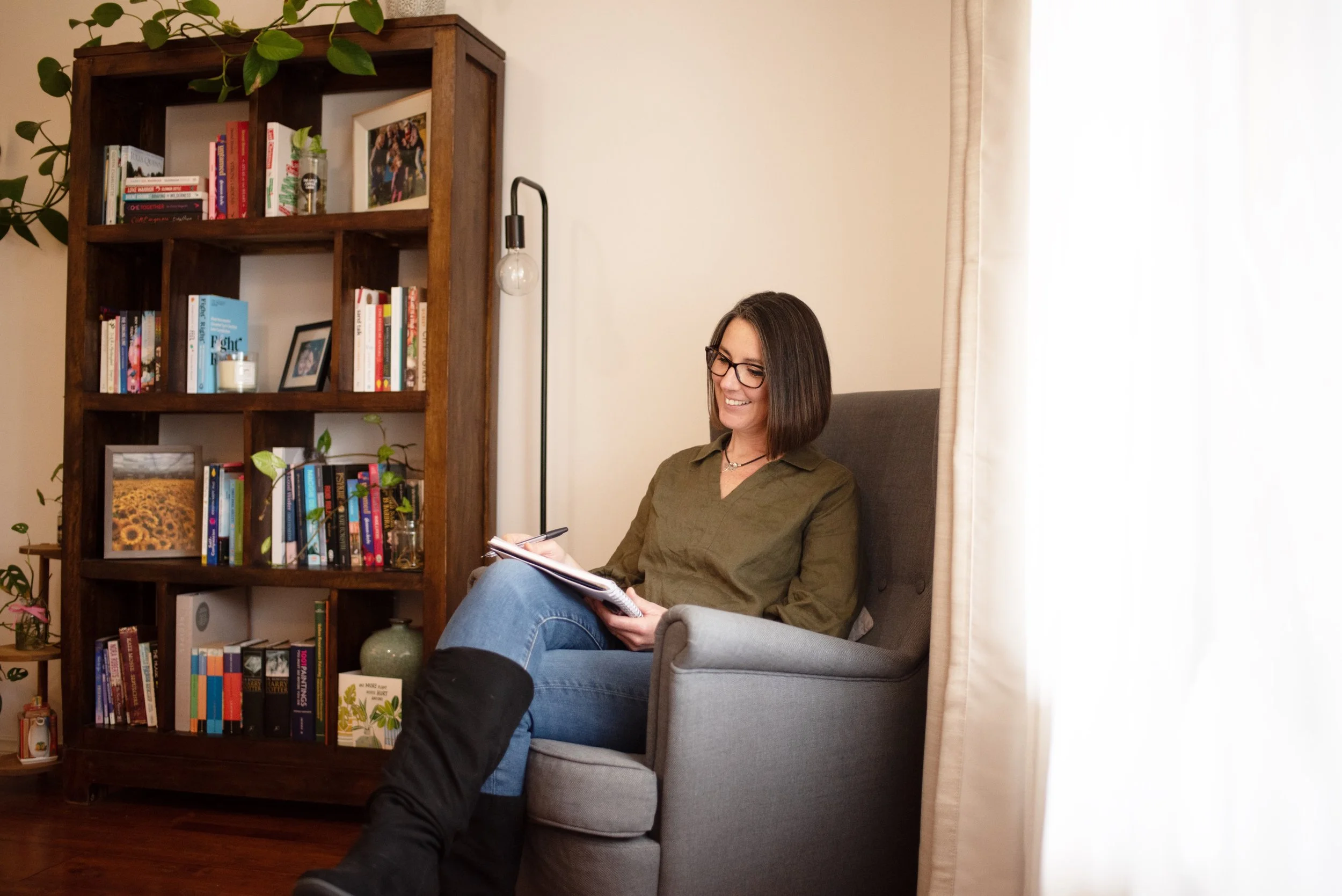 A woman with glasses reading a notebook while sitting on a grey armchair in a living room, next to a wooden bookshelf filled with books and decorative items, with a sheer beige curtain on the right side.