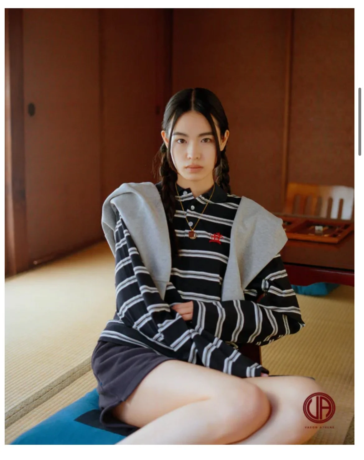 室内に座っている若い女性のポートレート。パーカーとストライプのシャツを着て、カジュアルな服装。背景は木製の壁と家具。