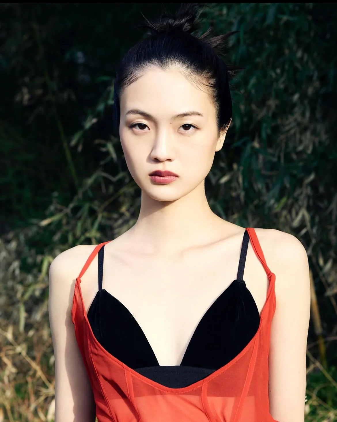 顔を向けている女性のポートレート。背景は緑の木々と葉。女性は赤と黒のドレスを着ている。