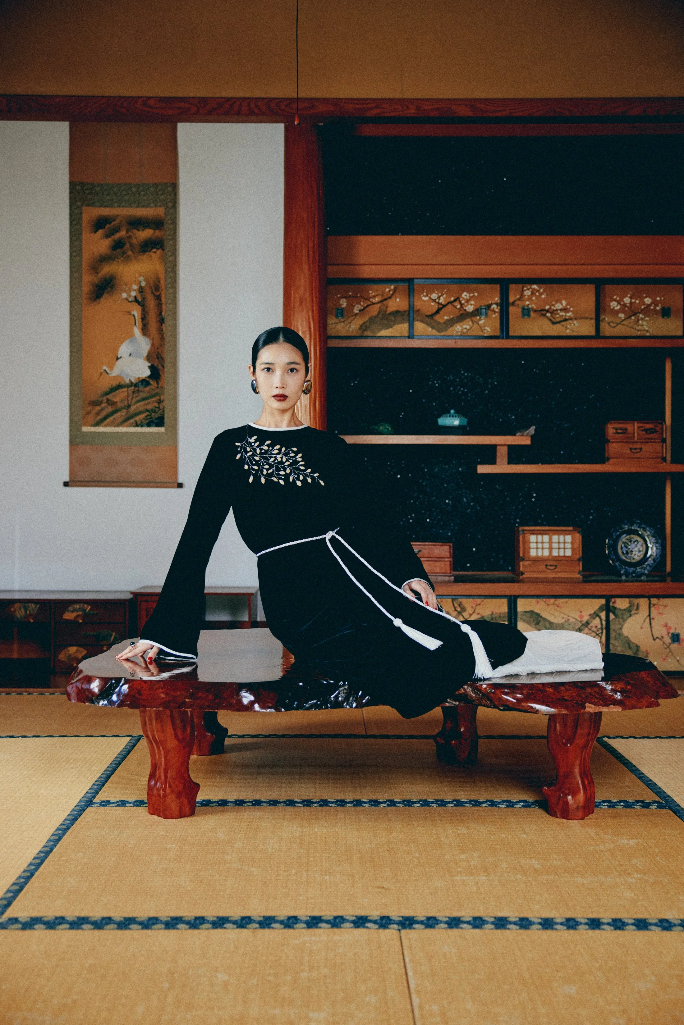 和室の床に座る女性。背景には伝統的な日本画や掛軸、木製の引き出しと棚。女性は黒い着物に白い刺繍と紐を身に着けている。