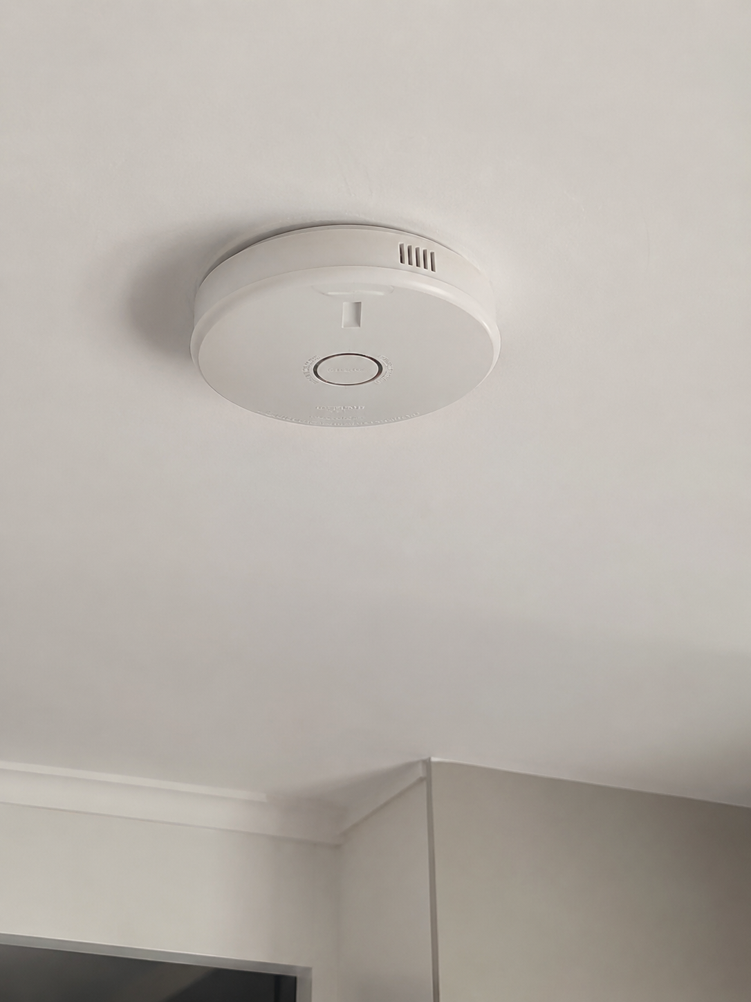 White smoke detector mounted on a white ceiling near a corner of a room.