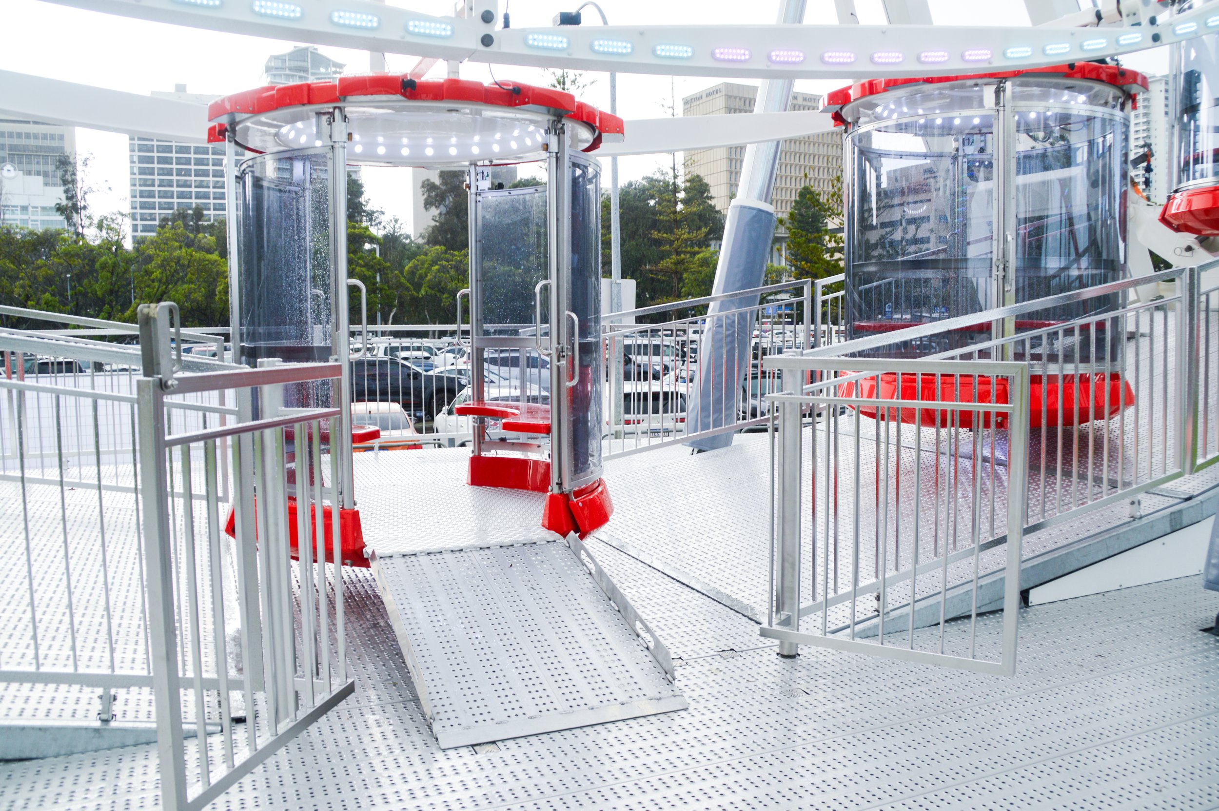A large outdoor Ferris wheel with enclosed gondolas, featuring metal railings and platforms for boarding, set against a cityscape with tall buildings and trees.