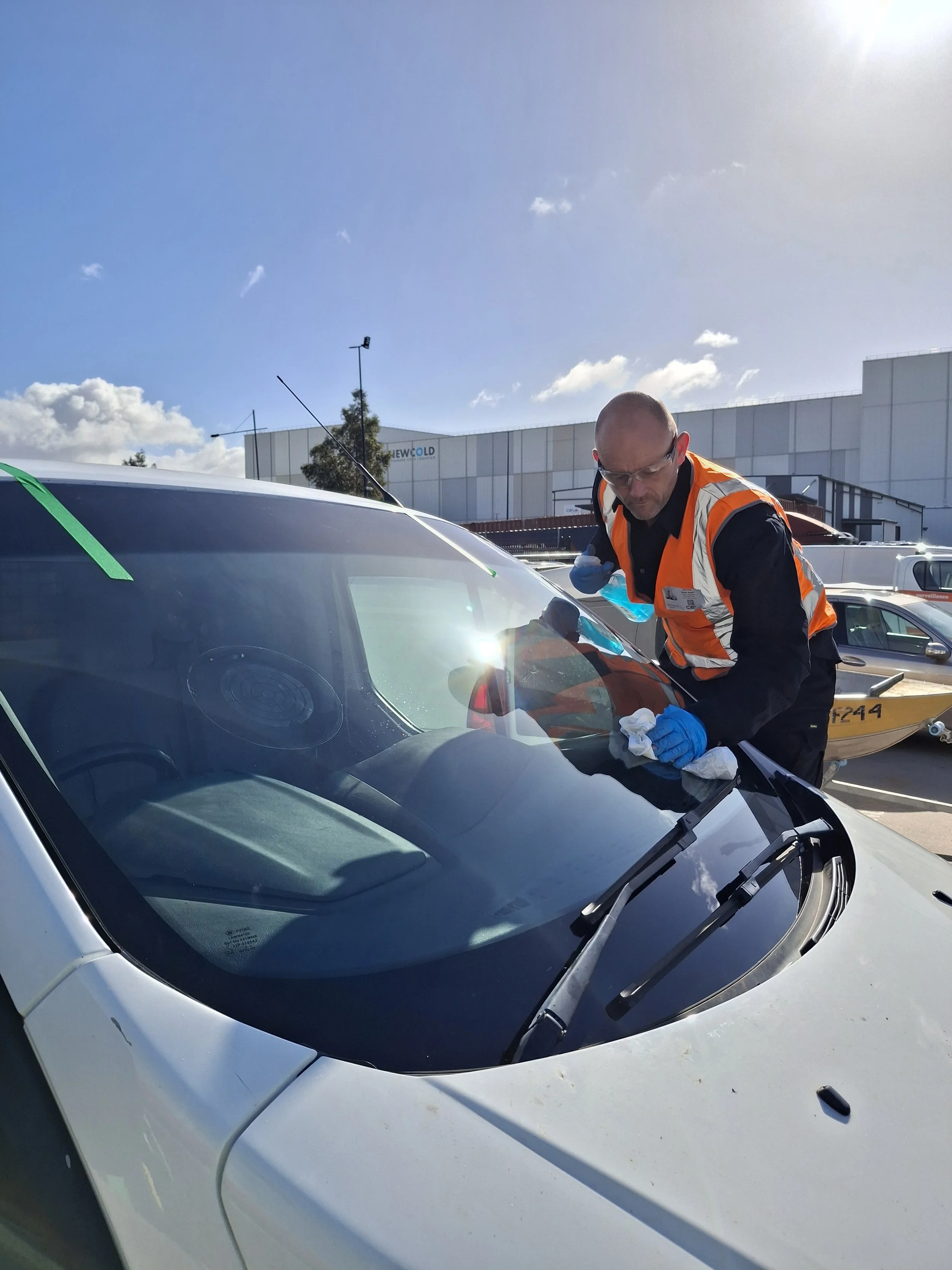 A man wearing safety glasses and an orange safety vest inspecting or cleaning a white car's windshield in a parking lot on a sunny day.
