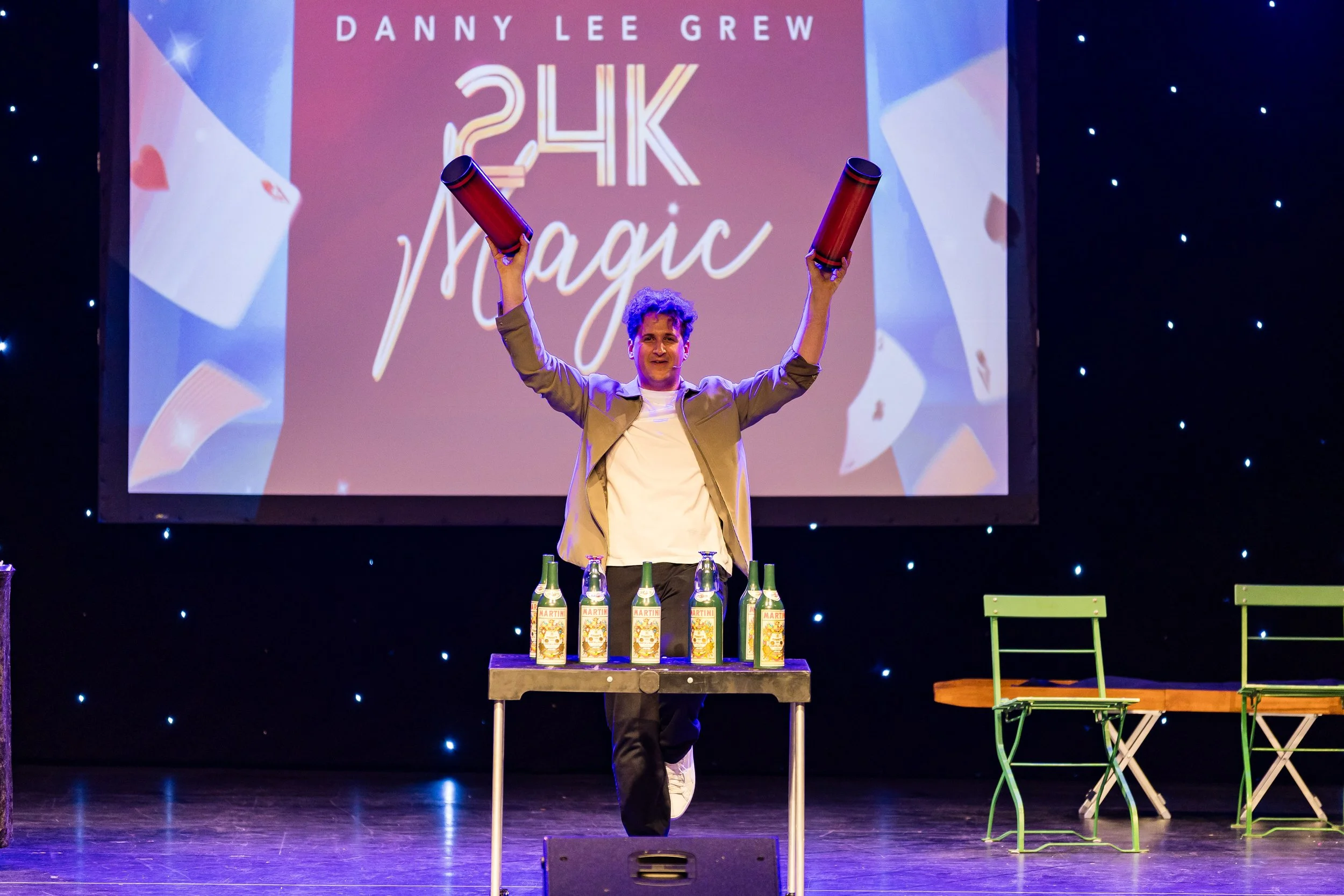 A man is standing on stage holding two red cylinders in the air with arms raised. There are six bottles of Martini sparkling drink on a table in front of him. The stage background displays the text 'DANNY LEE GREW 24K Magic' and has a decorated backd
