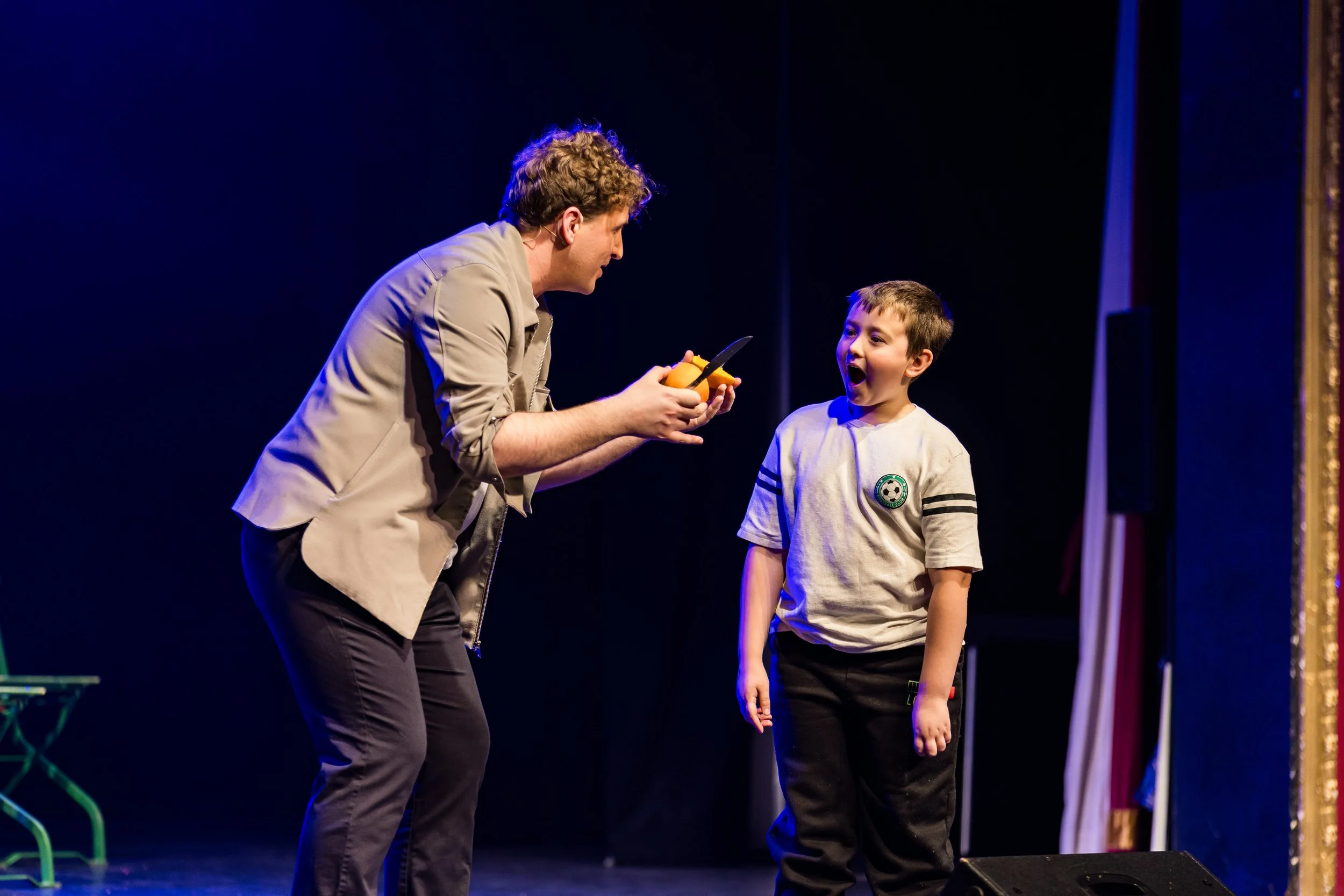 Man and boy on stage with dark background, man holding yellow object and knife, boy wearing beige shirt with a soccer emblem, both engaged in a performance.