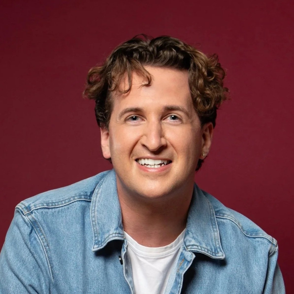 A smiling man with curly brown hair wearing a denim shirt and white t-shirt against a dark red background.