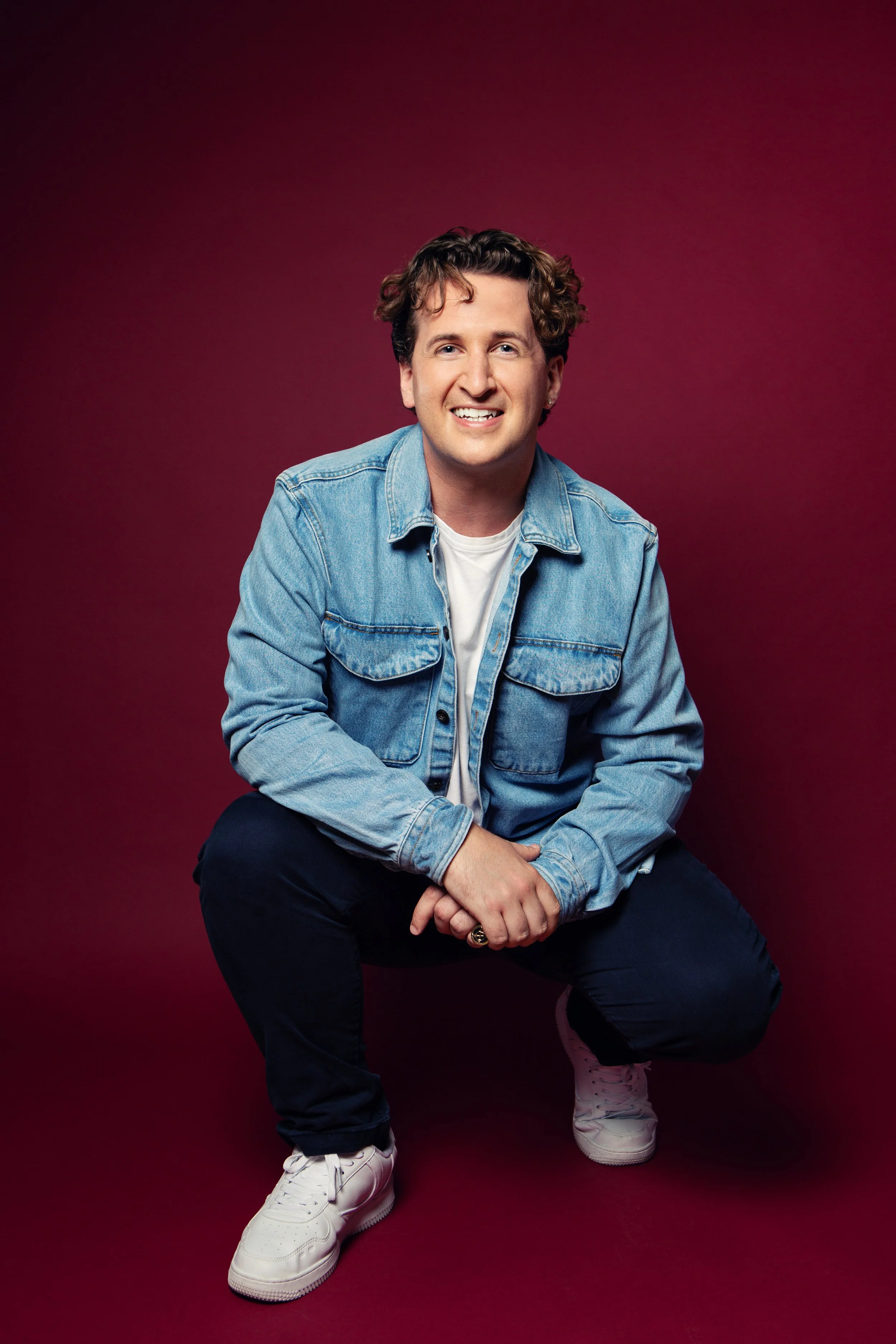A young man with curly hair wearing a denim jacket, white t-shirt, black pants, and white sneakers, sitting and smiling against a red background.
